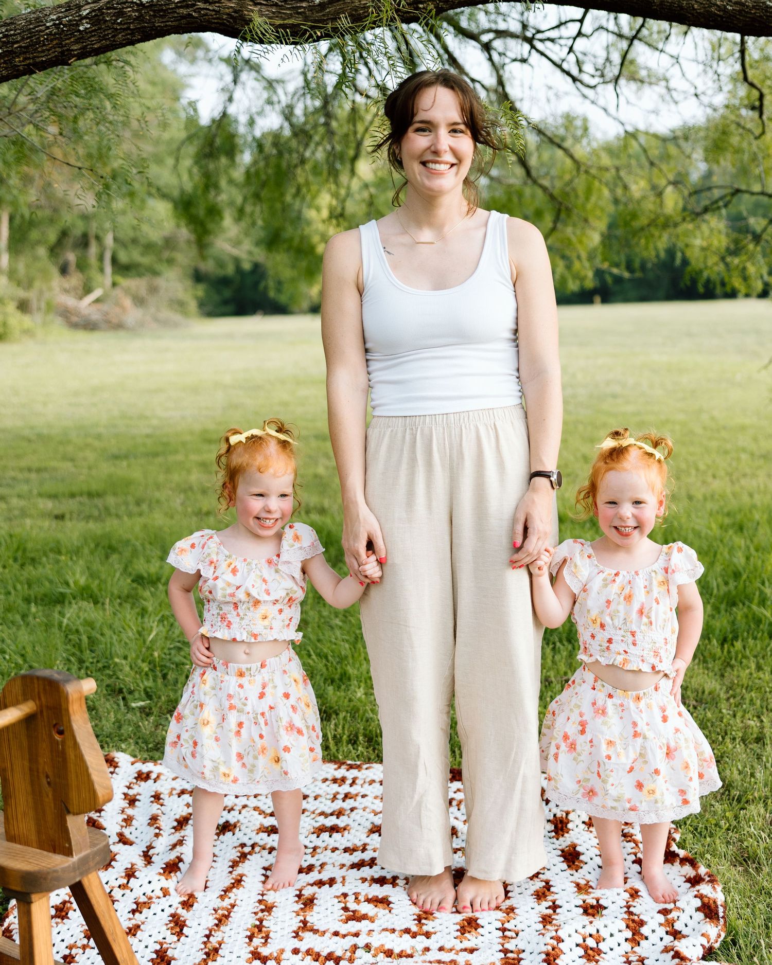 A smiling woman in beige pants holds hands with two red-haired twin girls in floral dresses on a grassy lawn.