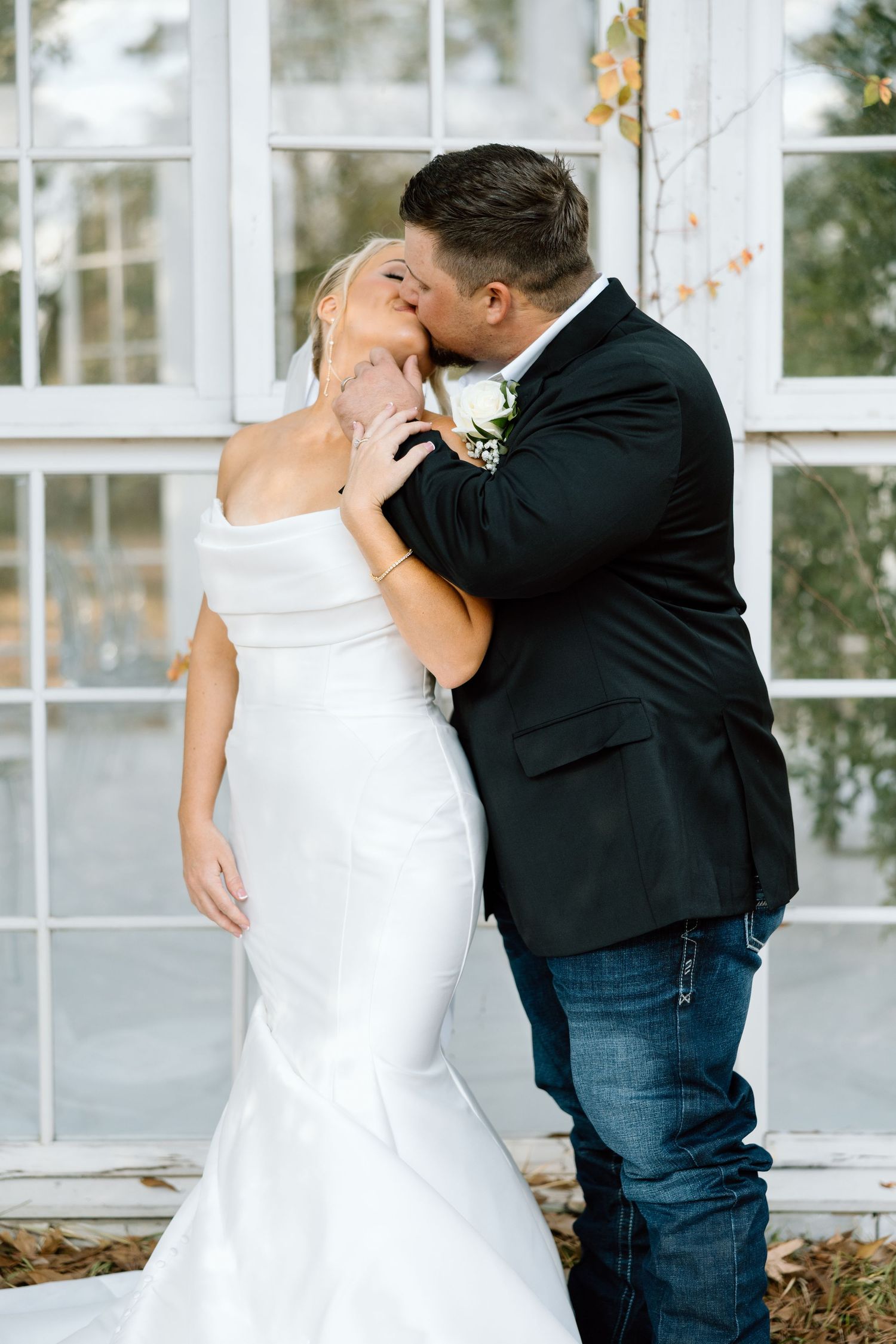 Bride and groom share a romantic kiss in front of white window panes, bride in fitted mermaid gown.