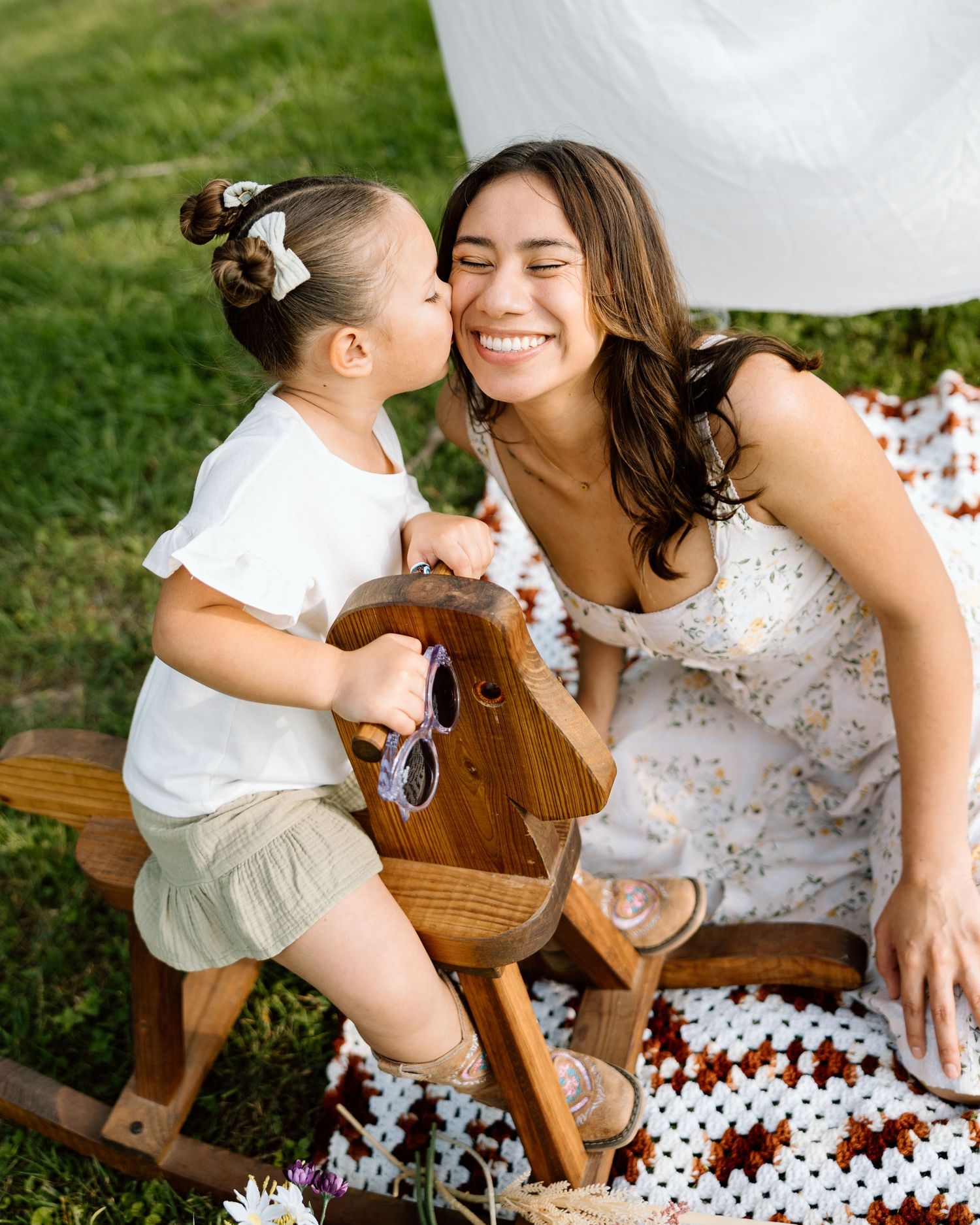 A toddler girl kisses her smiling mother on the cheek while sitting on a wooden rocking horse outdoors.