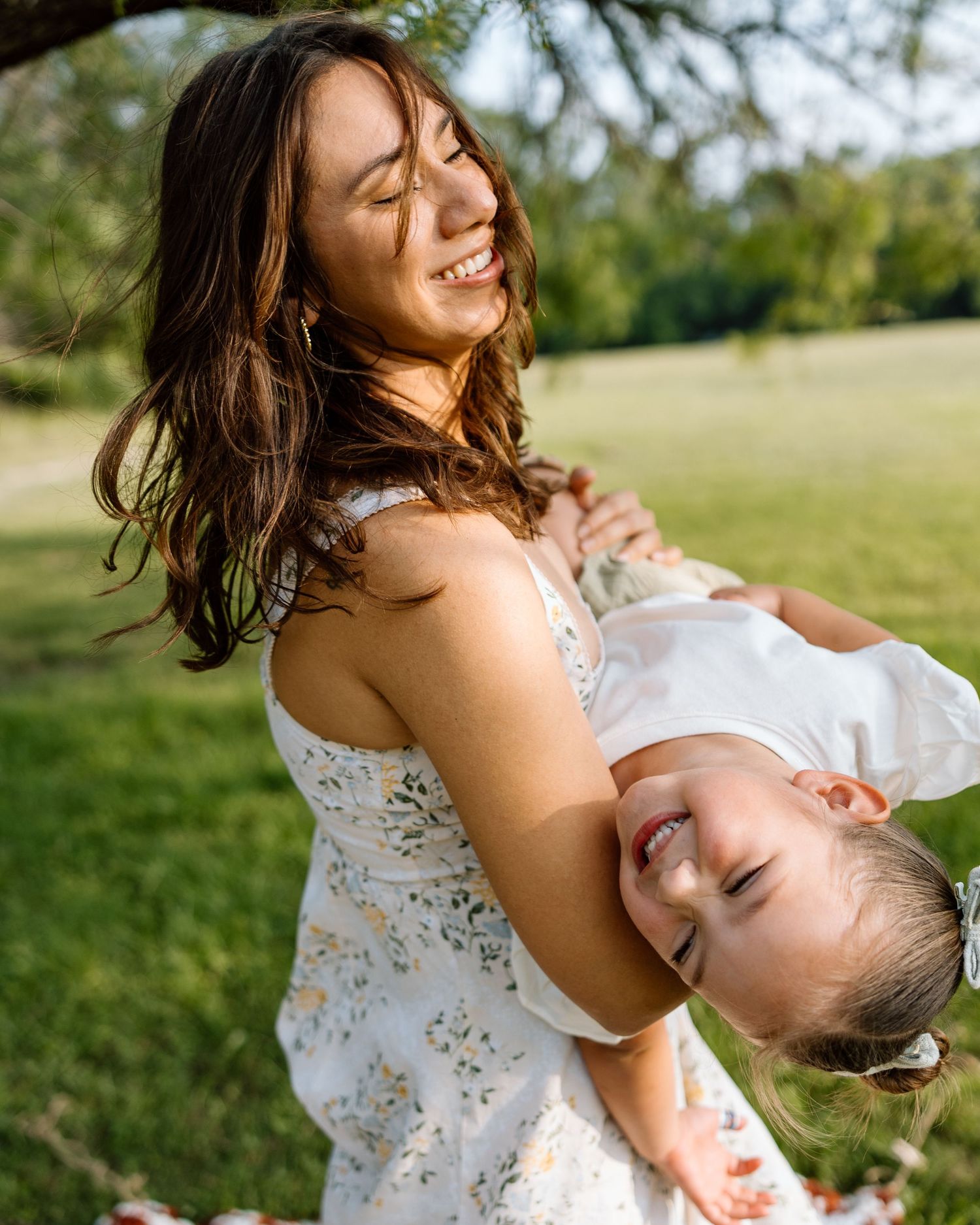 A joyful mother in a floral dress playfully holds her laughing toddler upside down in a sunny green park.