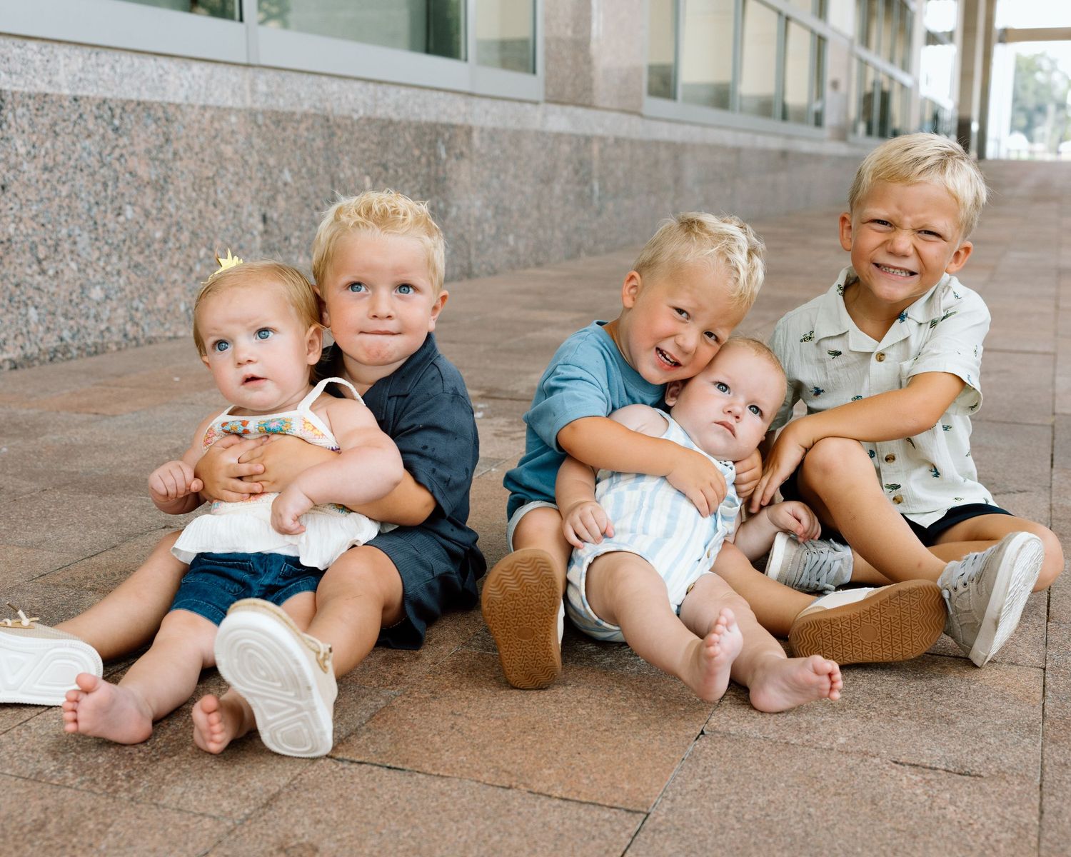 Six young toddler boys sitting together on outdoor steps, smiling and hugging in a candid family portrait.