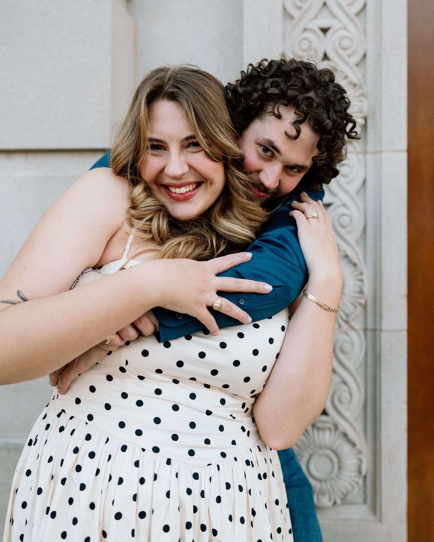 A smiling couple embraces warmly, woman in polka dot dress, man in teal shirt, posing against ornate white architecture.