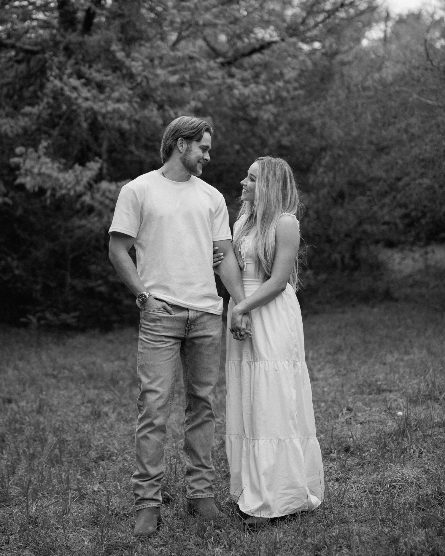 Black and white photo of a couple standing in a meadow, gazing lovingly at each other near tall trees.