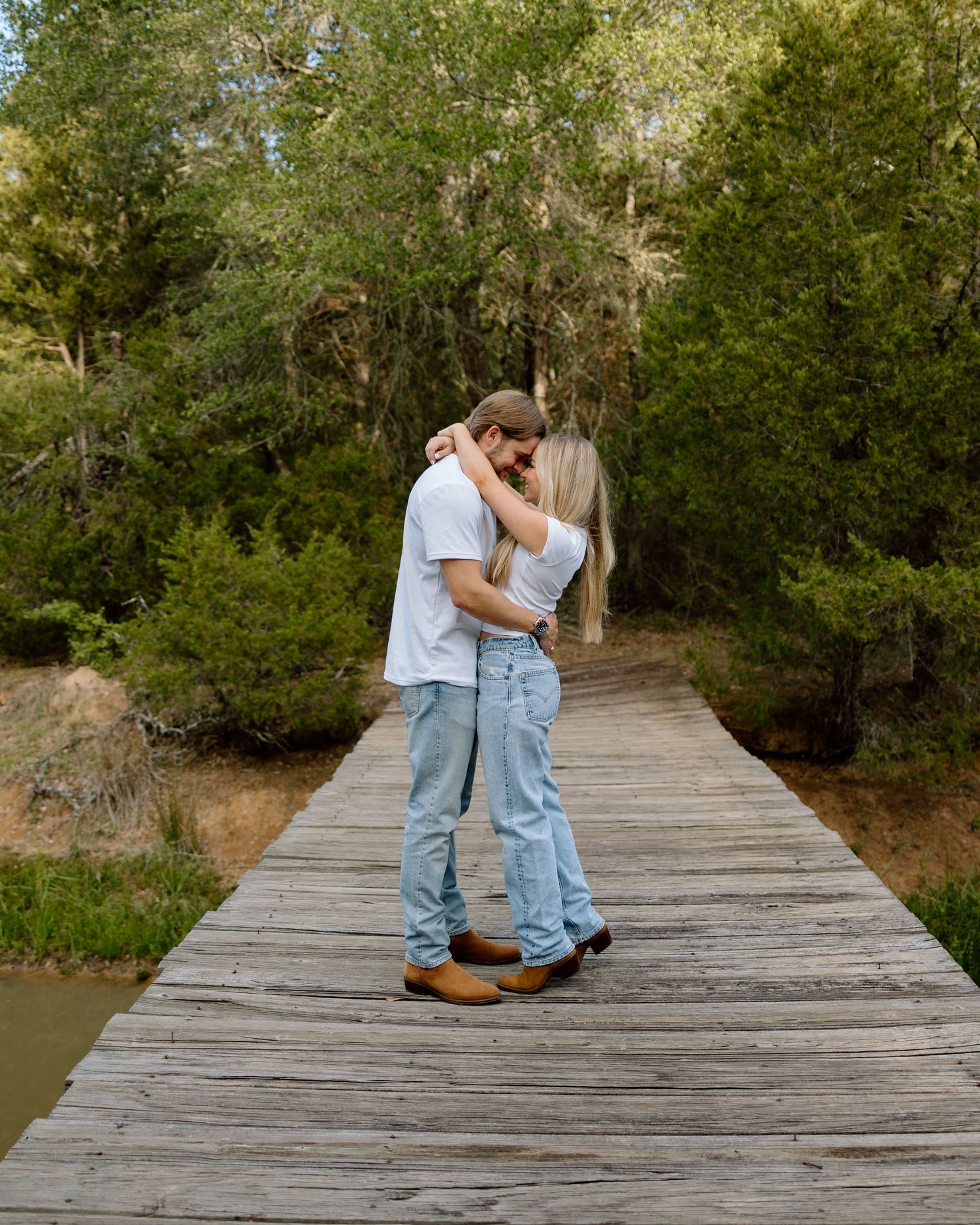Romantic couple embracing and kissing on a wooden bridge surrounded by lush green trees.