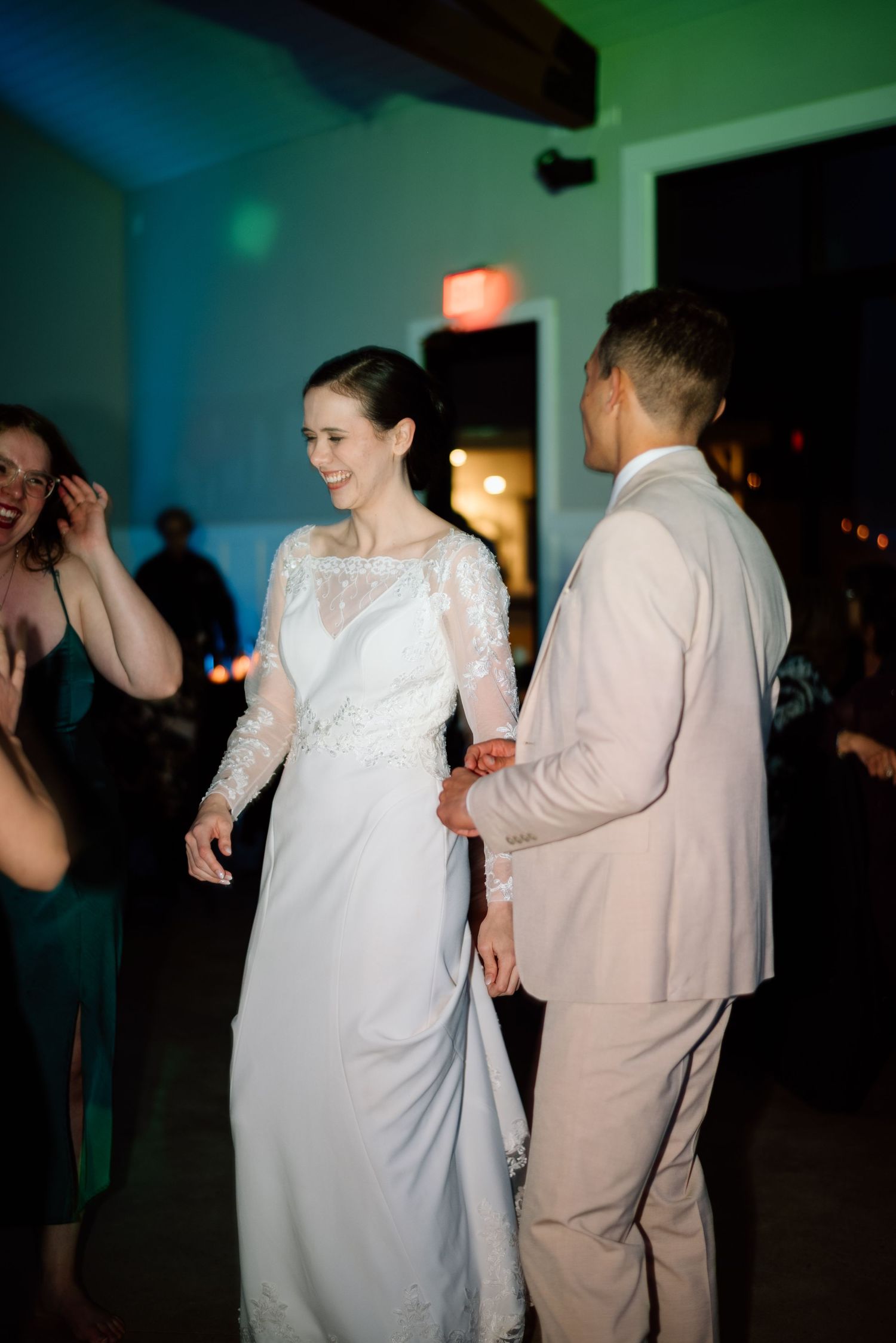Bride in lace wedding gown dances with groom in tan suit at indoor reception.