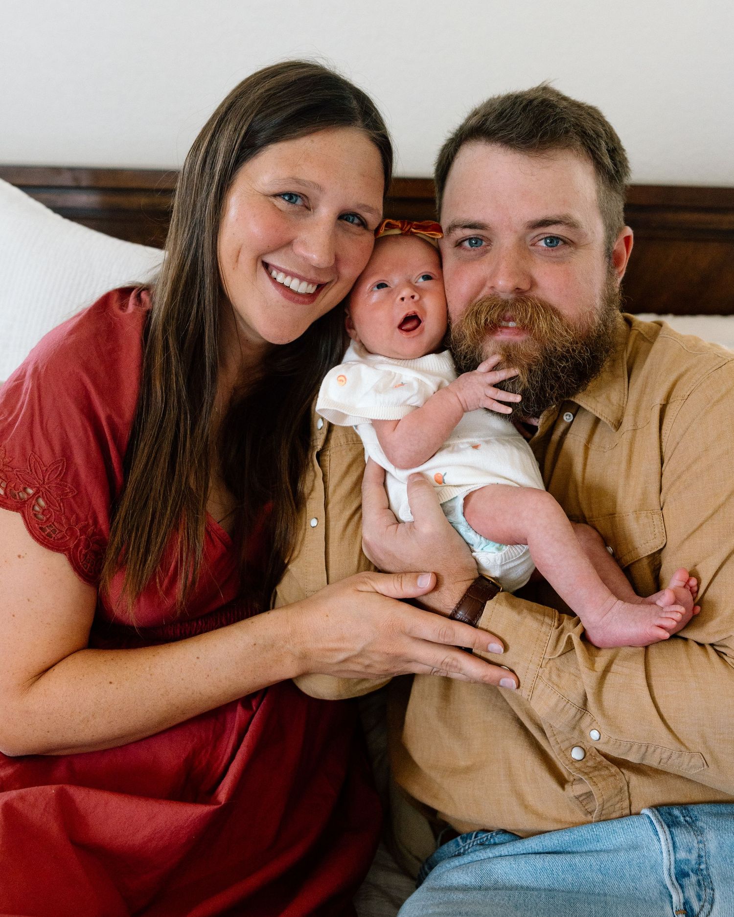 Happy couple smiling and holding their newborn baby while sitting together on a bed at home.