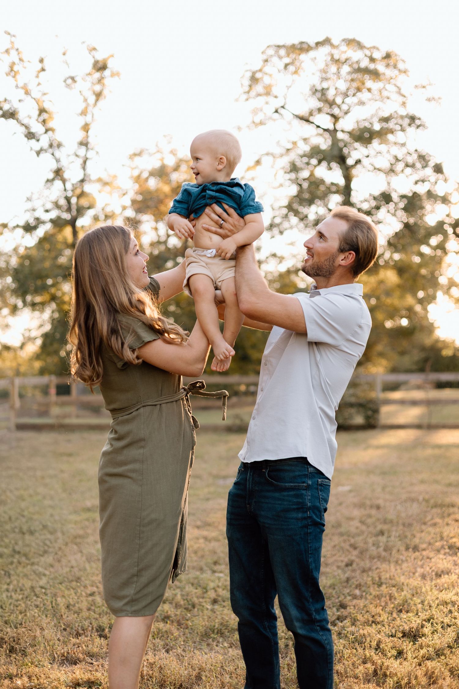 Happy young family with parents lifting smiling baby outdoors in golden hour sunlight near wooden fence.