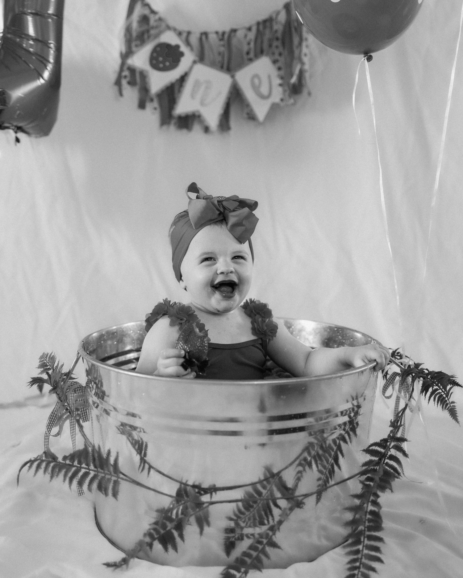Black and white photo of a laughing baby in a metal tub with ferns, wearing a bow headband at a first birthday.
