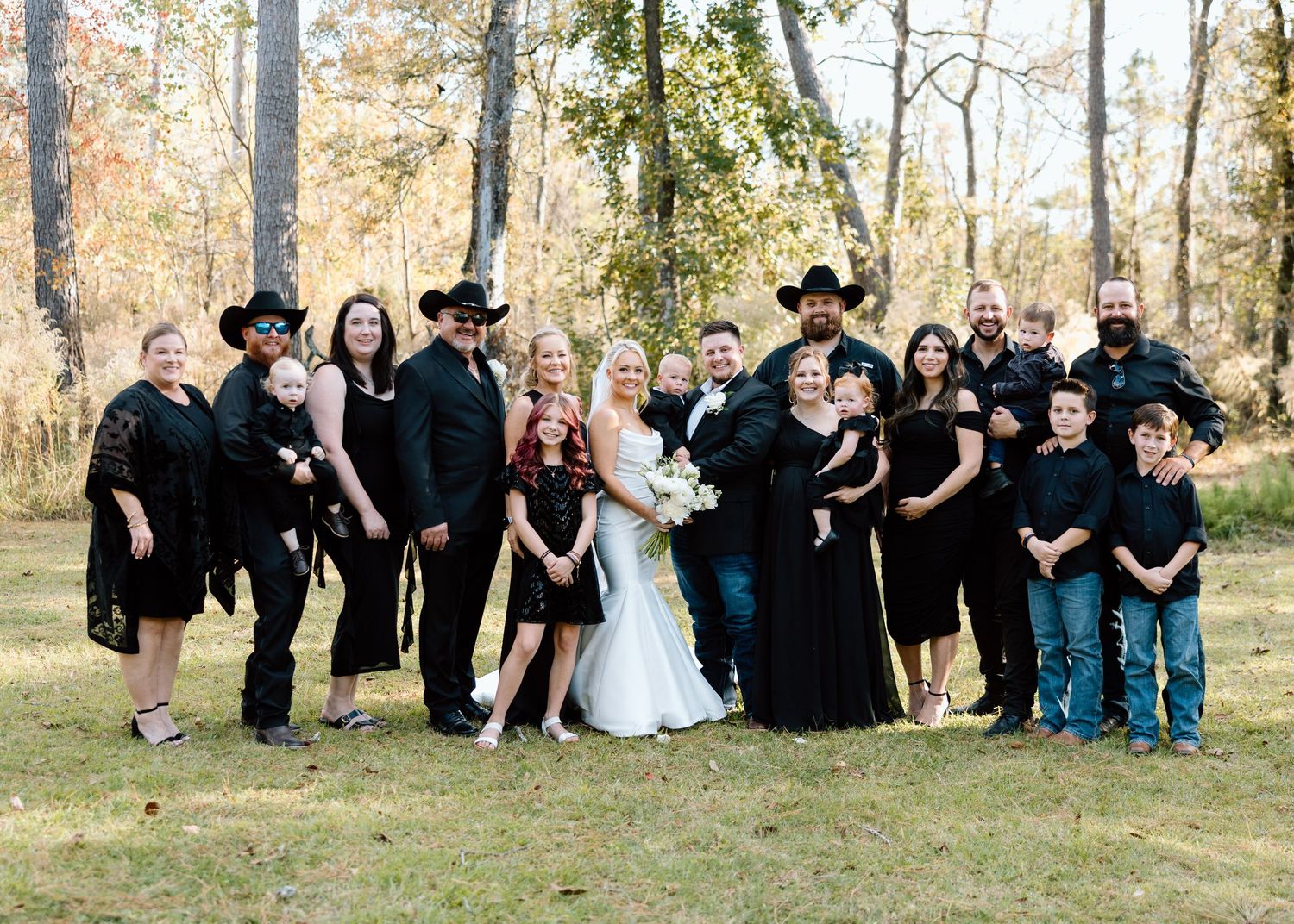 Wedding party in black attire poses with bride and groom on grassy lawn surrounded by autumn trees.