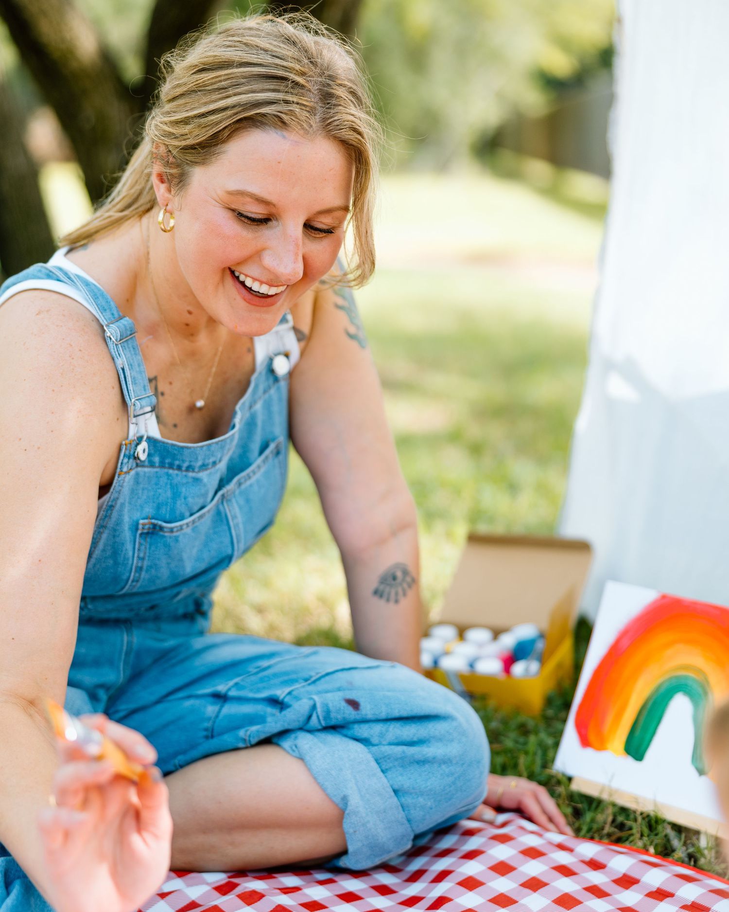 Woman in denim overalls smiling while painting a rainbow canvas outdoors on a red checkered blanket.