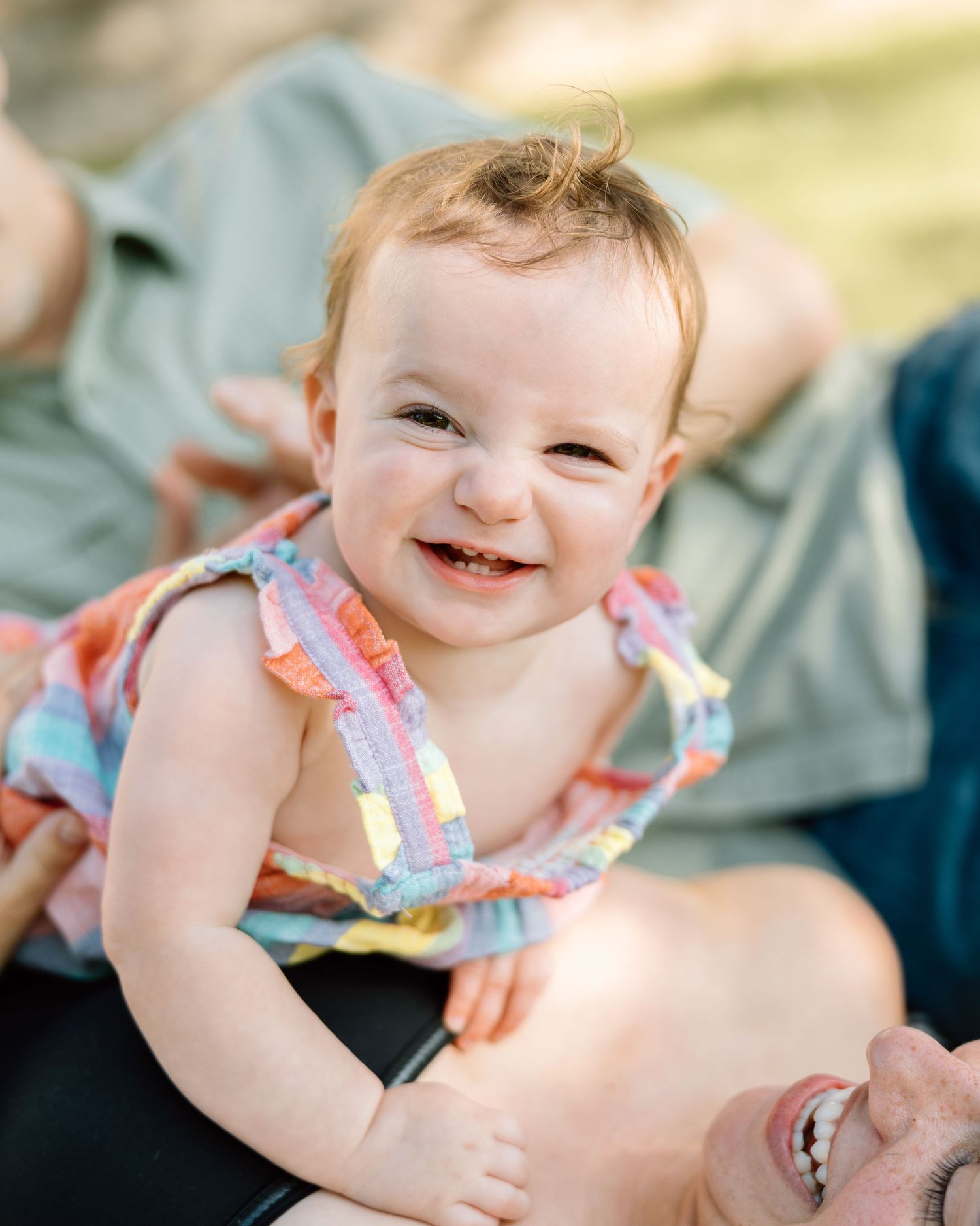 Happy smiling baby girl in rainbow dress laughing while being held by parent outdoors.