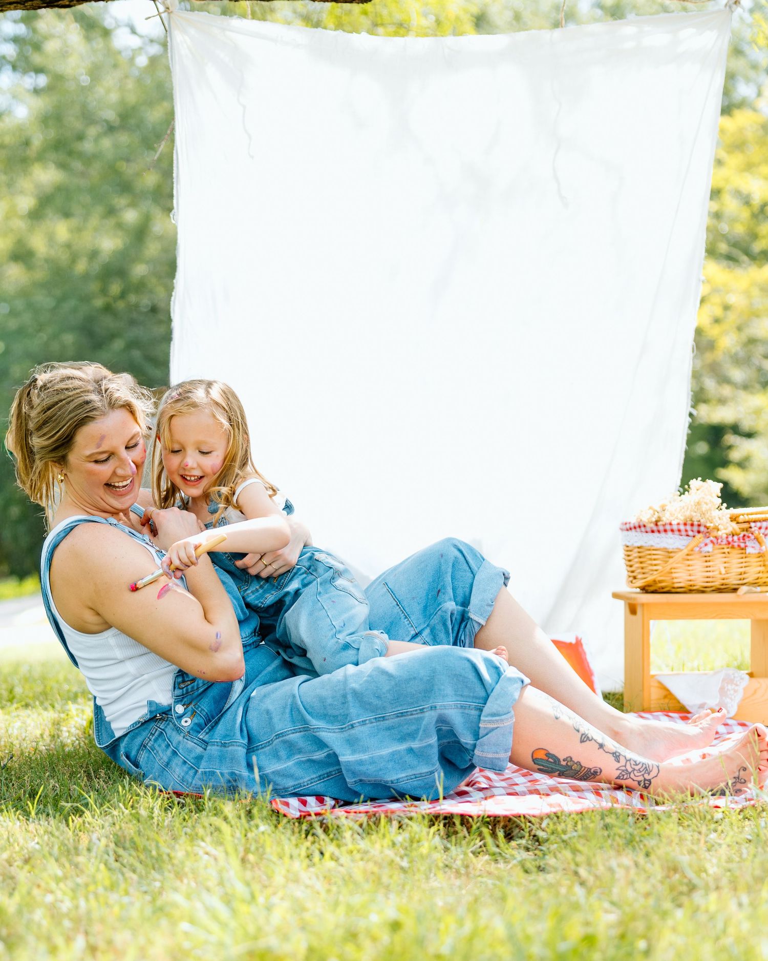 A mother and daughter laughing together on a picnic blanket outdoors with a white sheet backdrop.