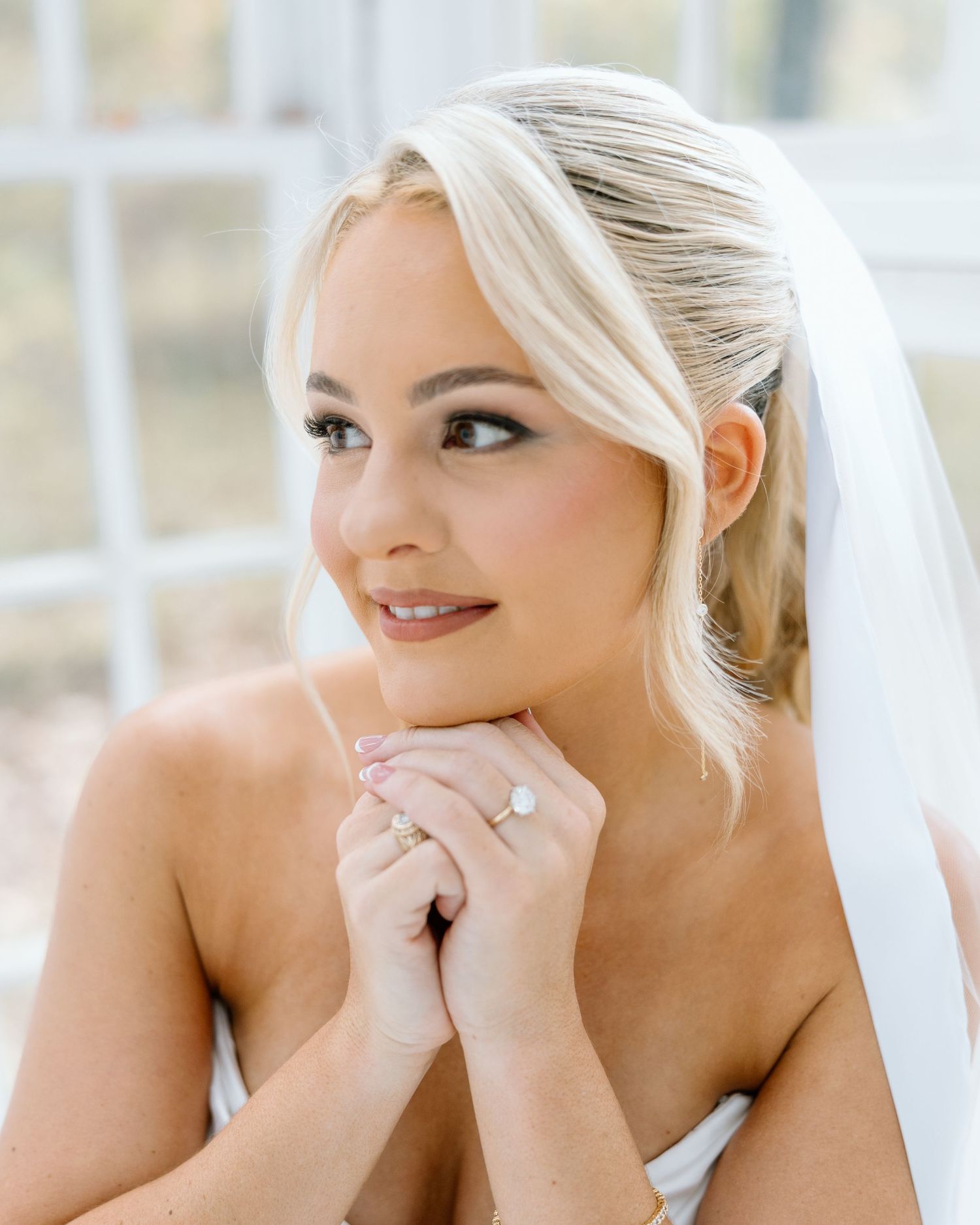Blonde bride wearing a white veil and engagement ring poses with hands clasped, showcasing elegant bridal makeup.