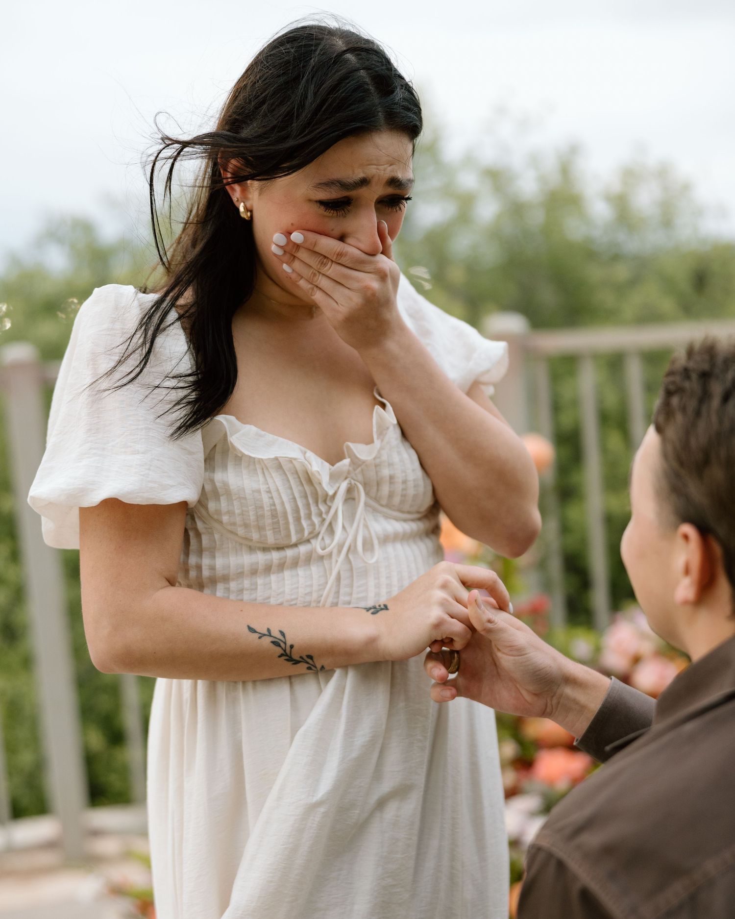Emotional woman in white dress covering her mouth during a marriage proposal outdoors.