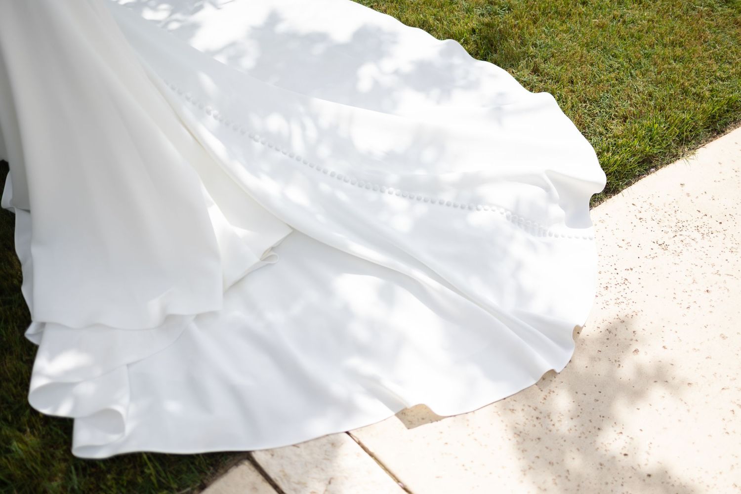Close-up detail of a flowing white wedding dress train against grass and concrete.