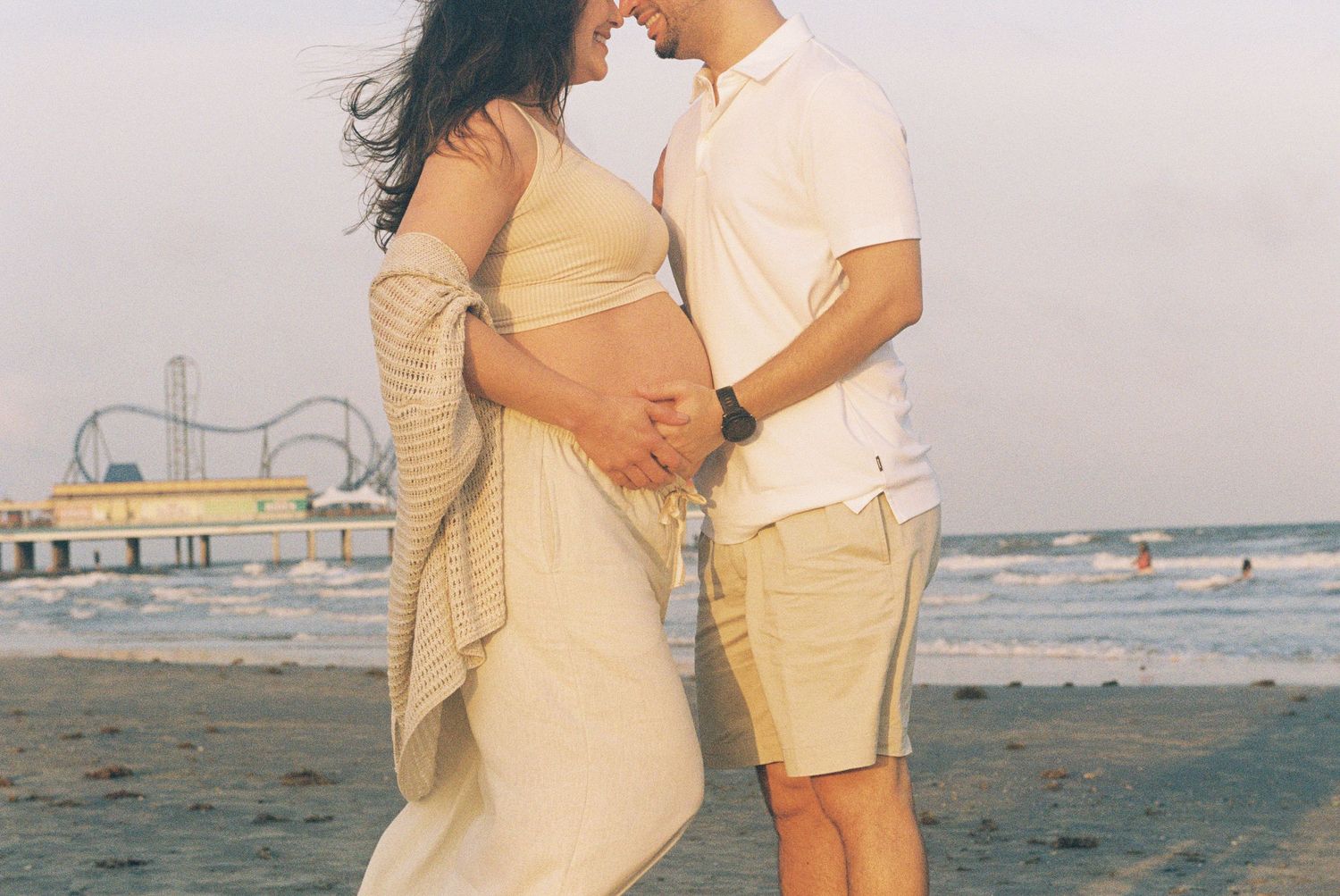 A couple embraces on the beach at sunset with an amusement park pier in the background.