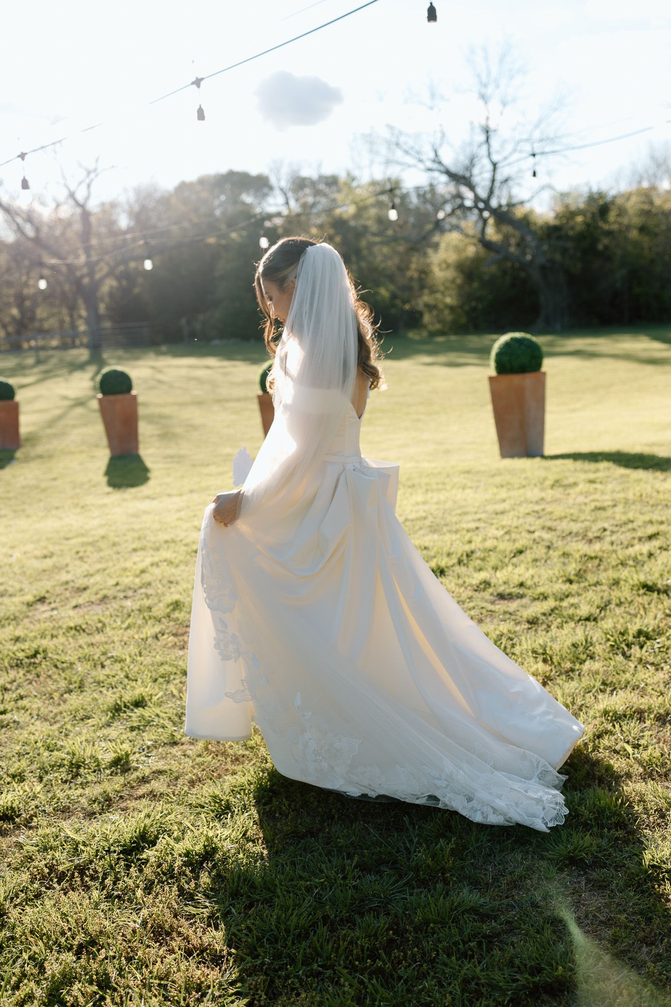 Bride in flowing white dress stands on sunlit lawn during golden hour wedding photoshoot.