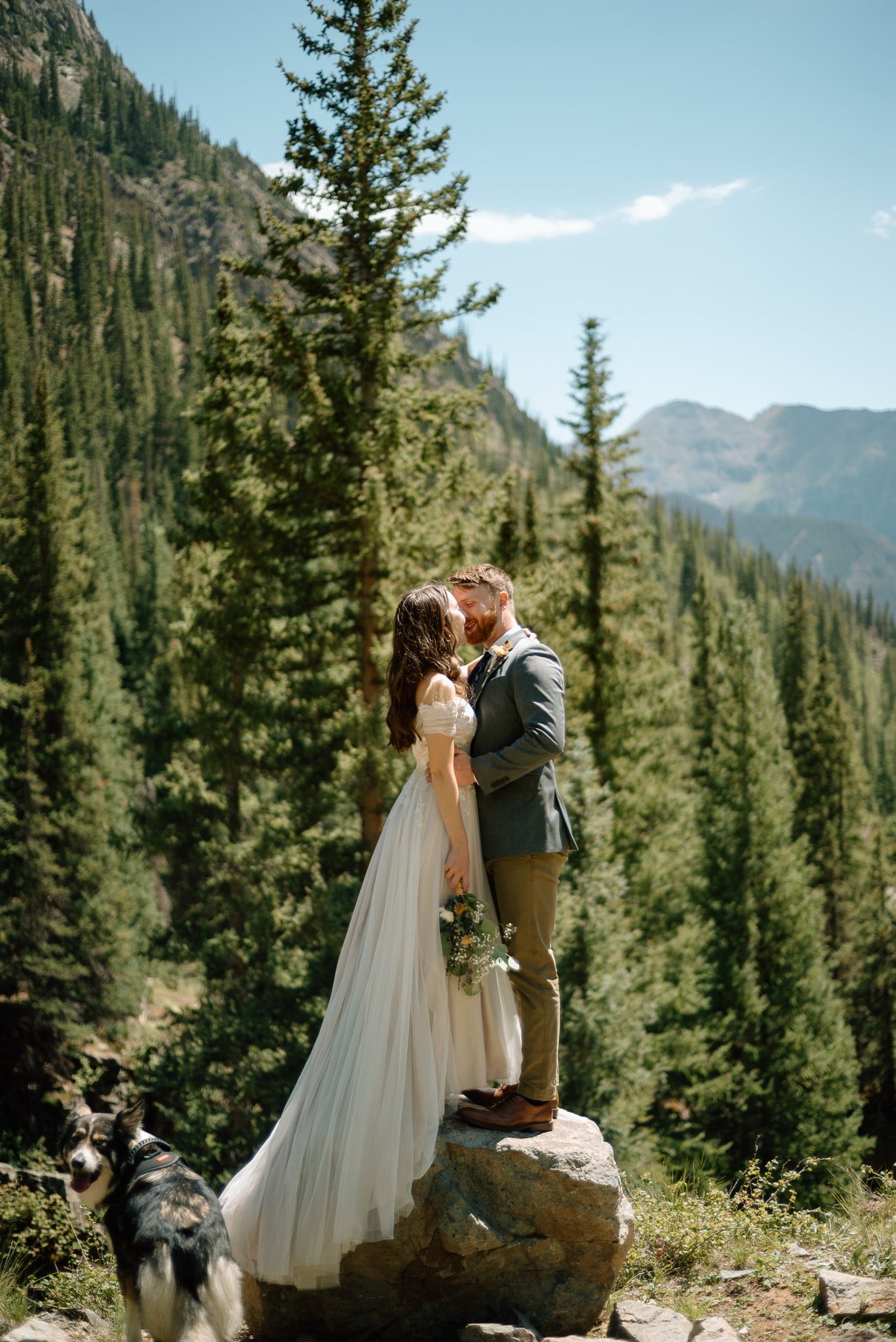 Couple shares romantic moment on mountain rock with dog watching below.