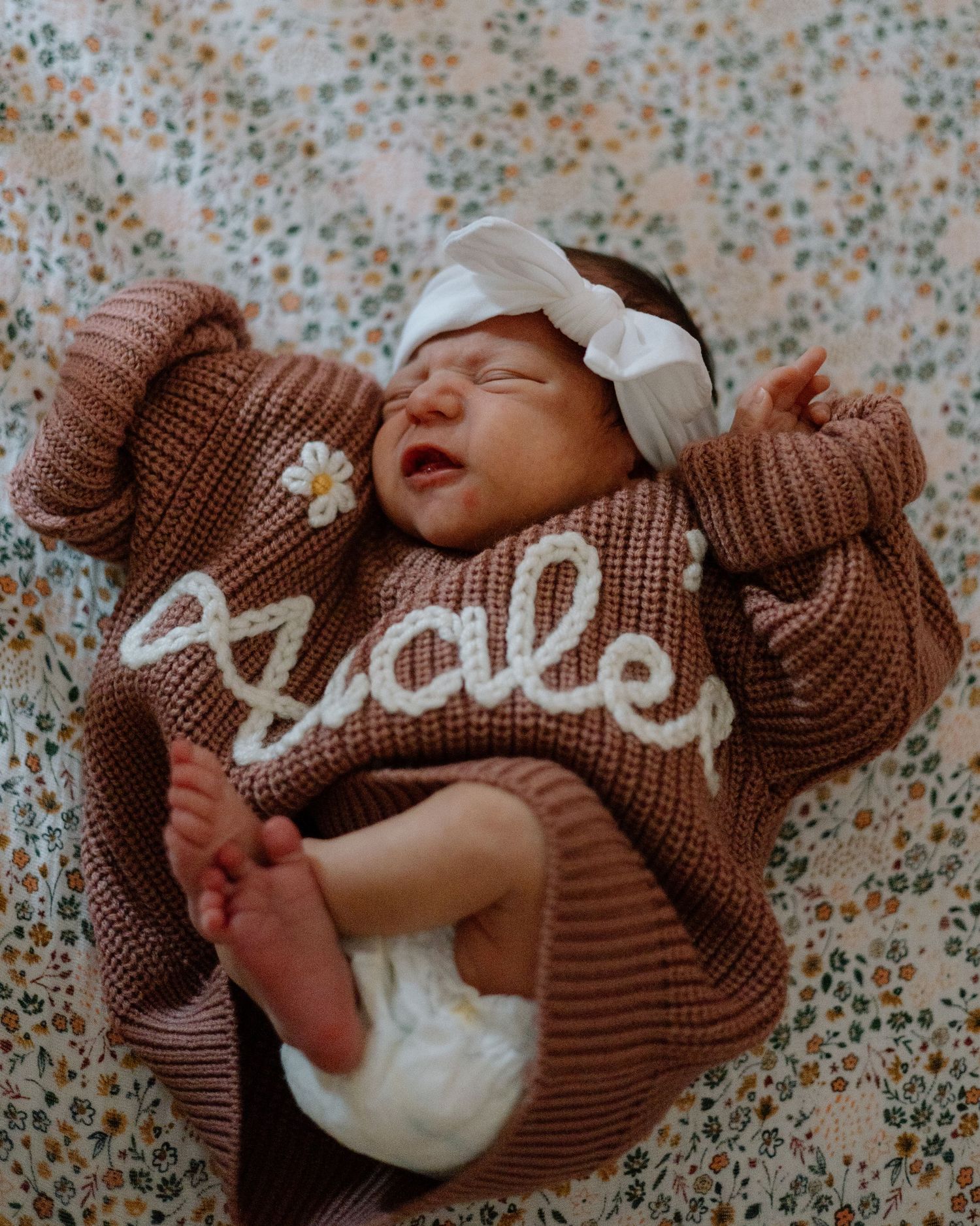 Newborn baby wearing brown knit sweater with bow headband laying on floral background.