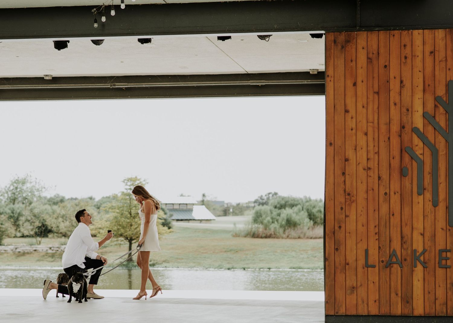 A tender proposal moment unfolds under an architectural overhang by the water.