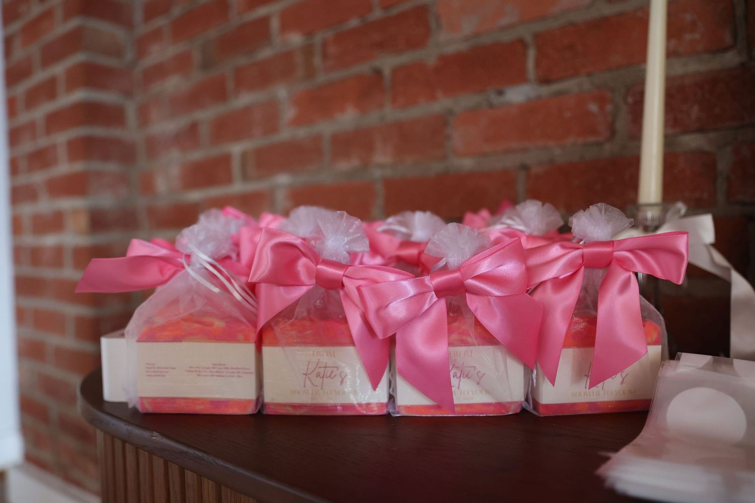 Row of pink favor boxes with bows arranged on a wooden surface against a brick wall.