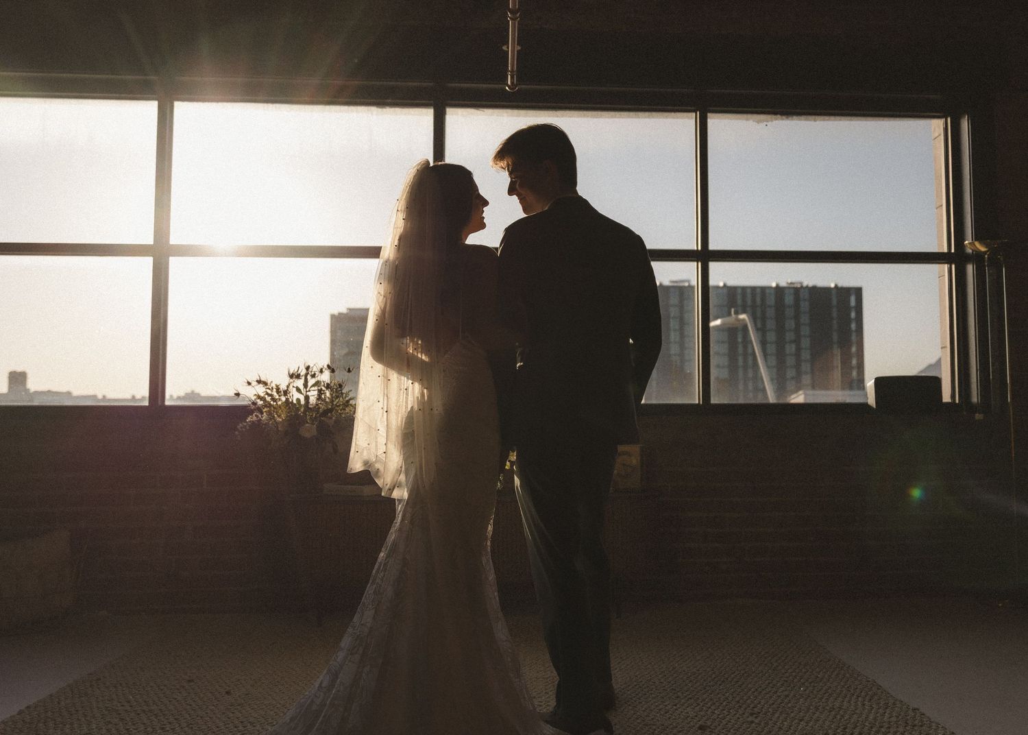 A romantic silhouette of a wedding couple standing against large windows with sunlight streaming in.