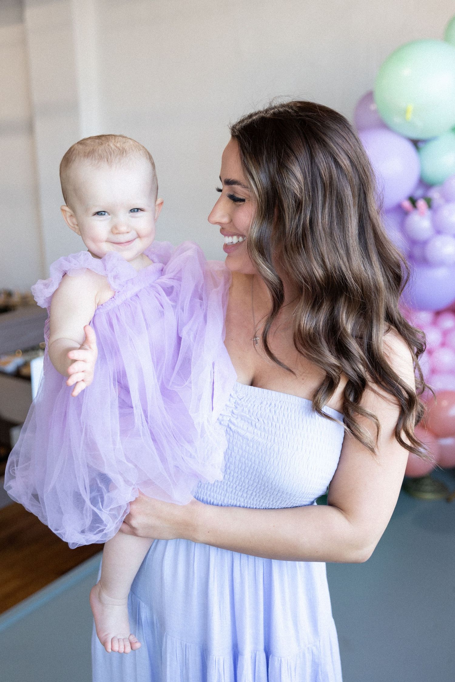 Woman in light blue dress holding baby wearing purple tulle dress with balloon decorations.