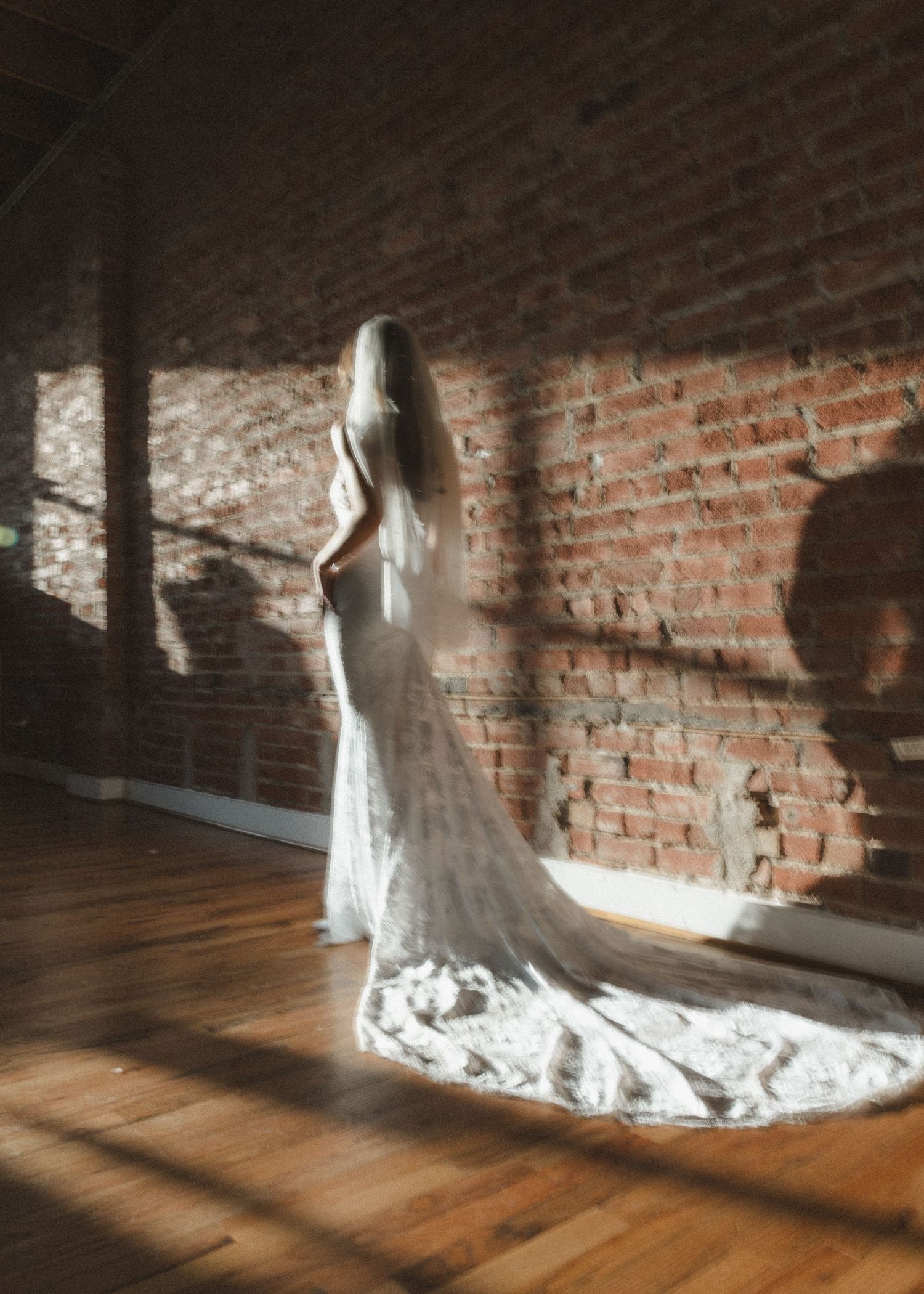 Long-trained lace wedding dress displayed against exposed brick wall with dramatic lighting.