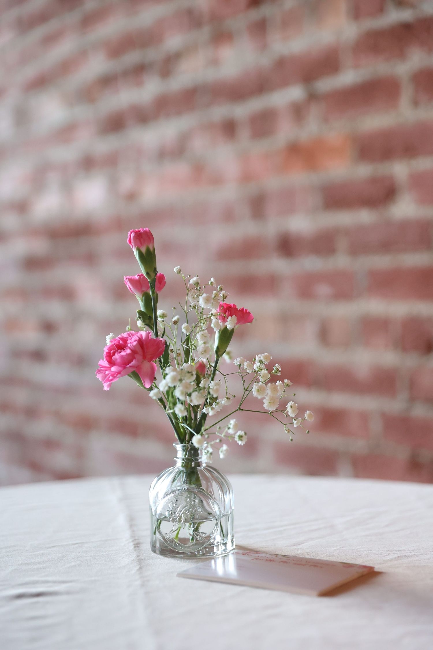 Small glass vase with pink carnations and baby's breath against rustic brick wall.