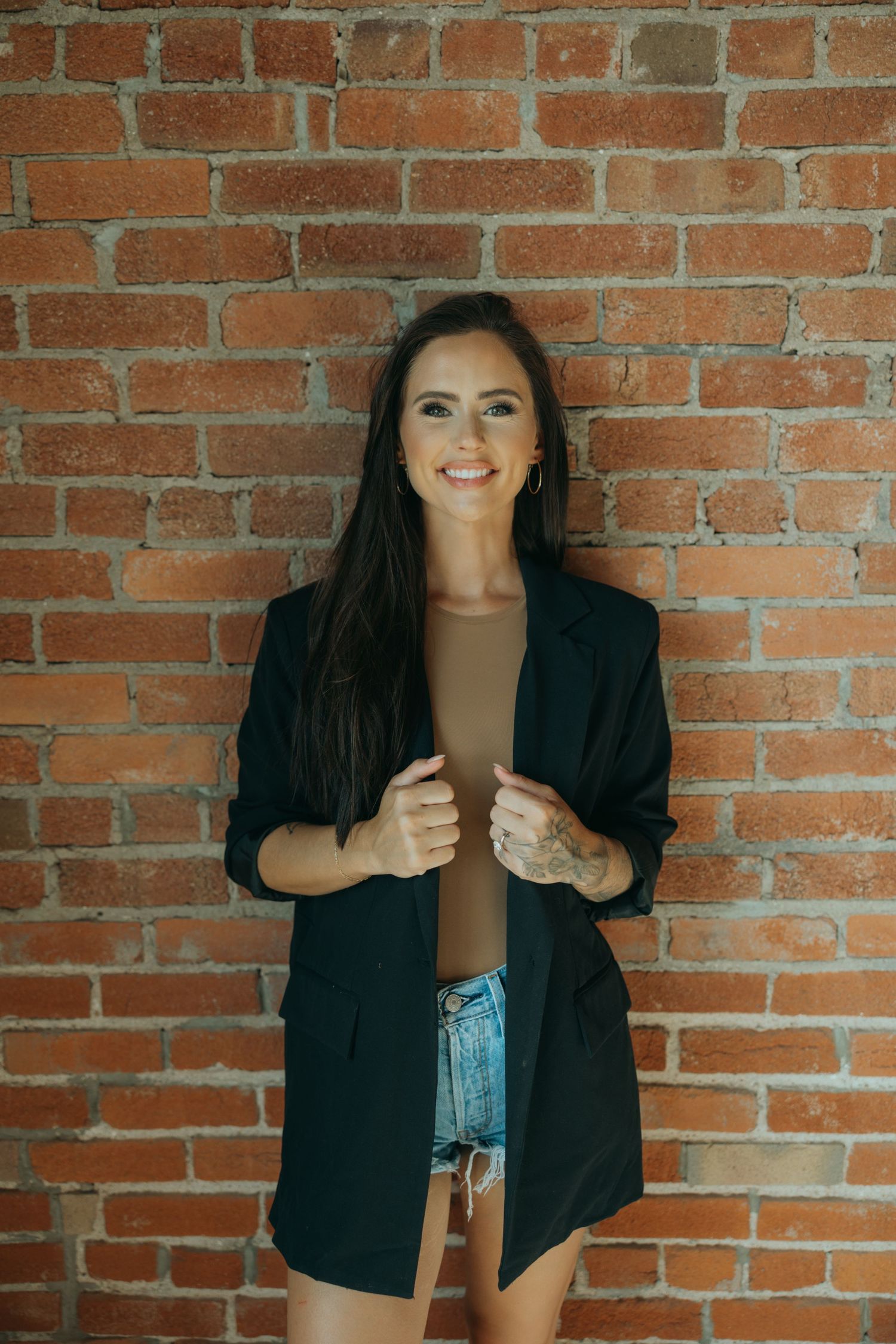 Professional portrait against exposed brick wall featuring a black blazer and denim shorts in an urban style.