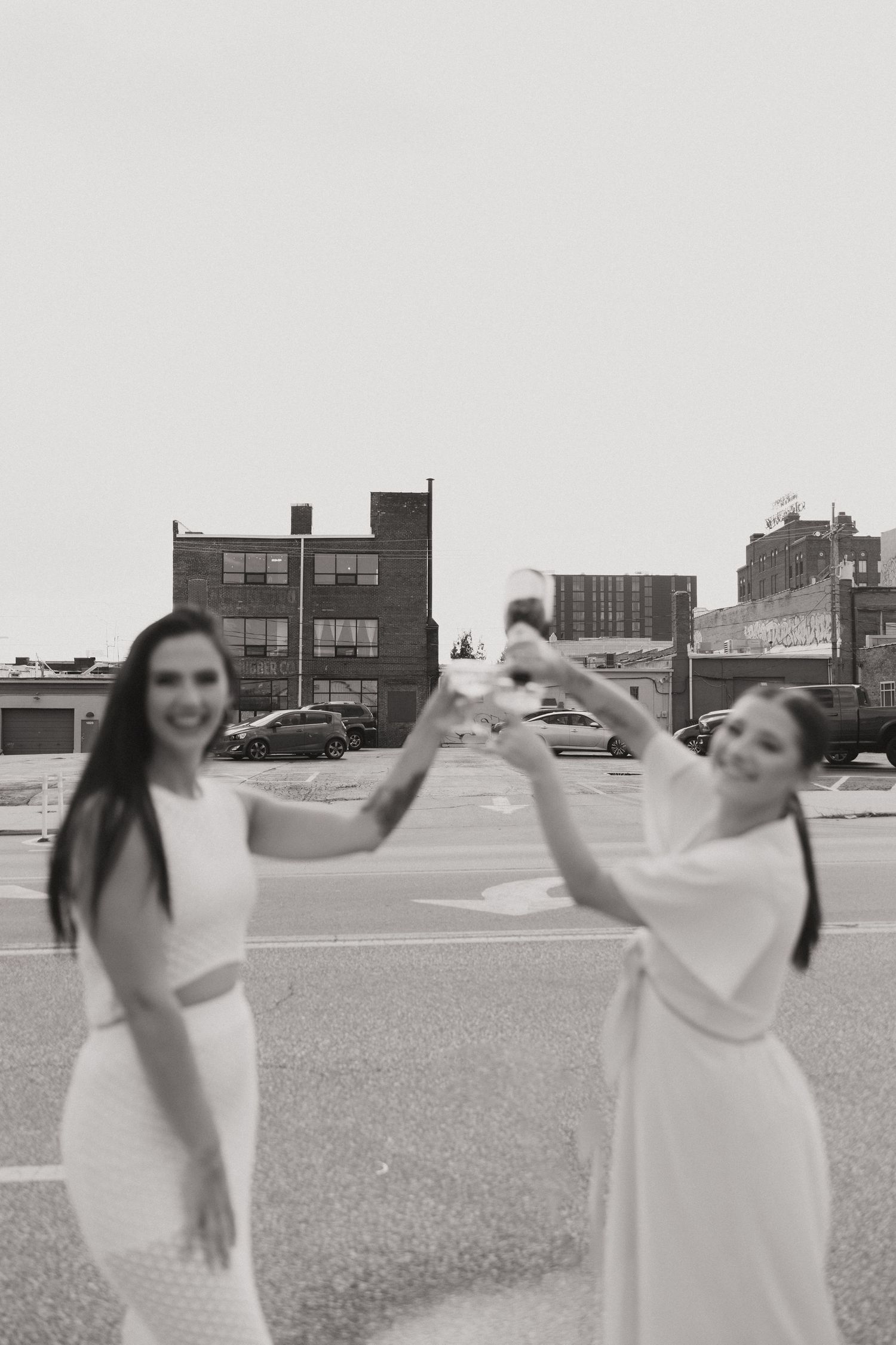 Black and white photo of two people dancing in a parking lot wearing light-colored dresses.