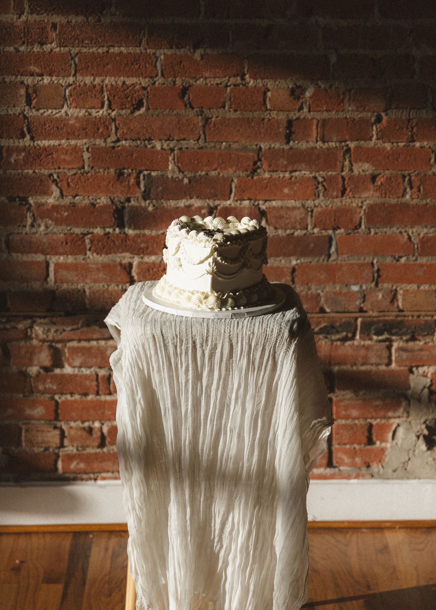 Wedding cake displayed atop draped fabric against rustic brick wall in natural lighting.