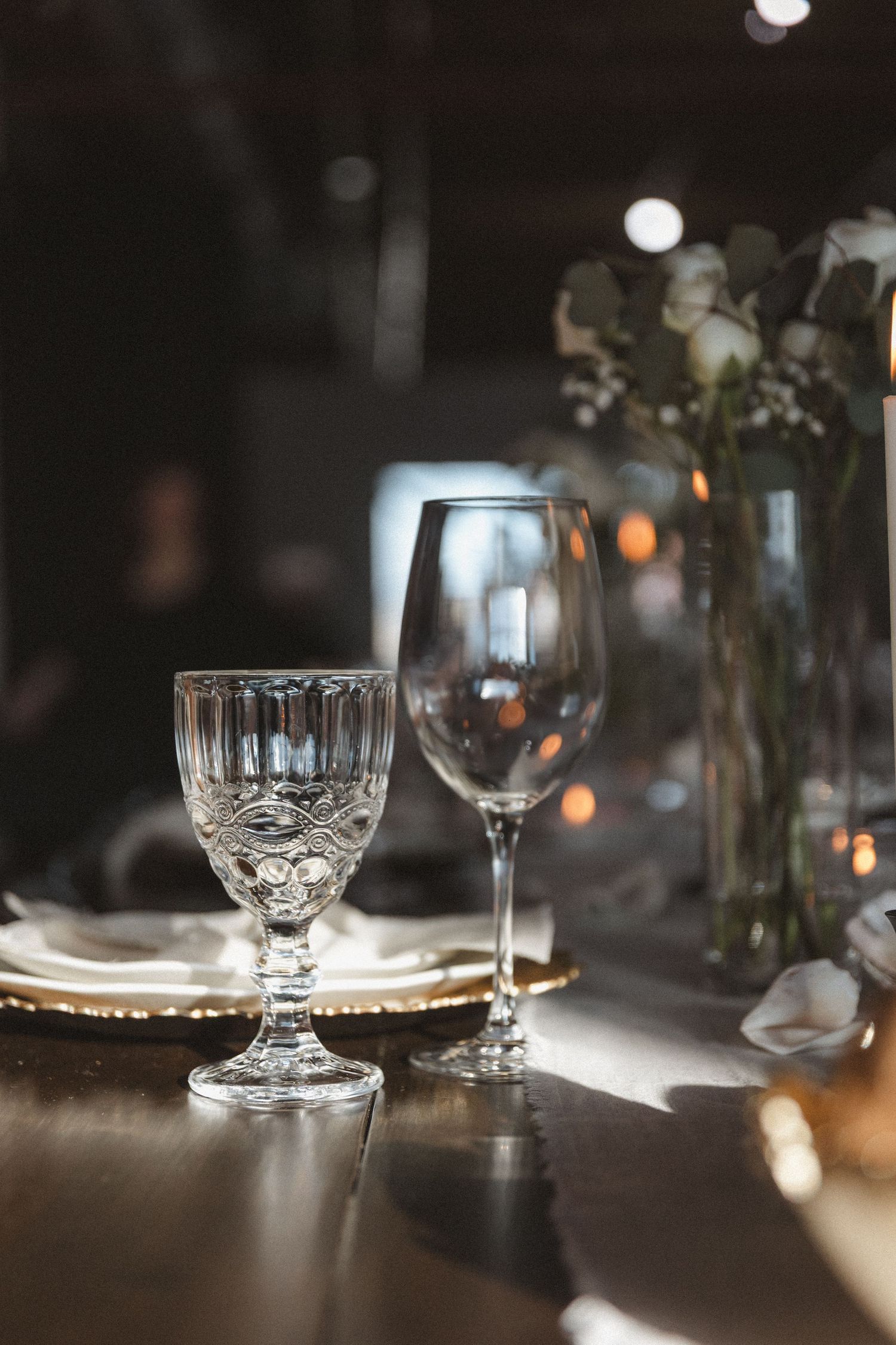 Crystal wine glasses on dining table with moody lighting and floral arrangements.