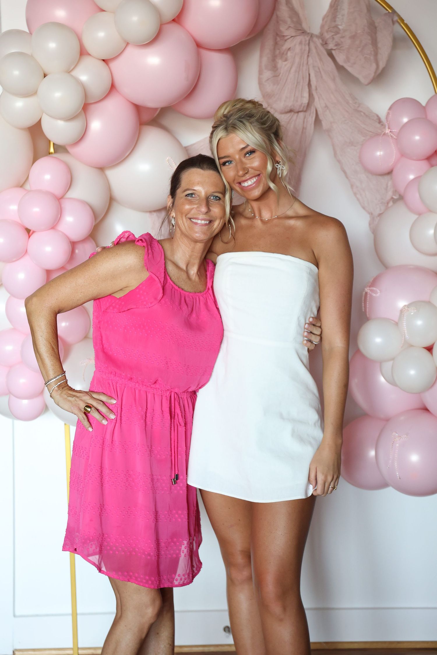 Two friends pose together in front of a pink balloon arch at a celebration.