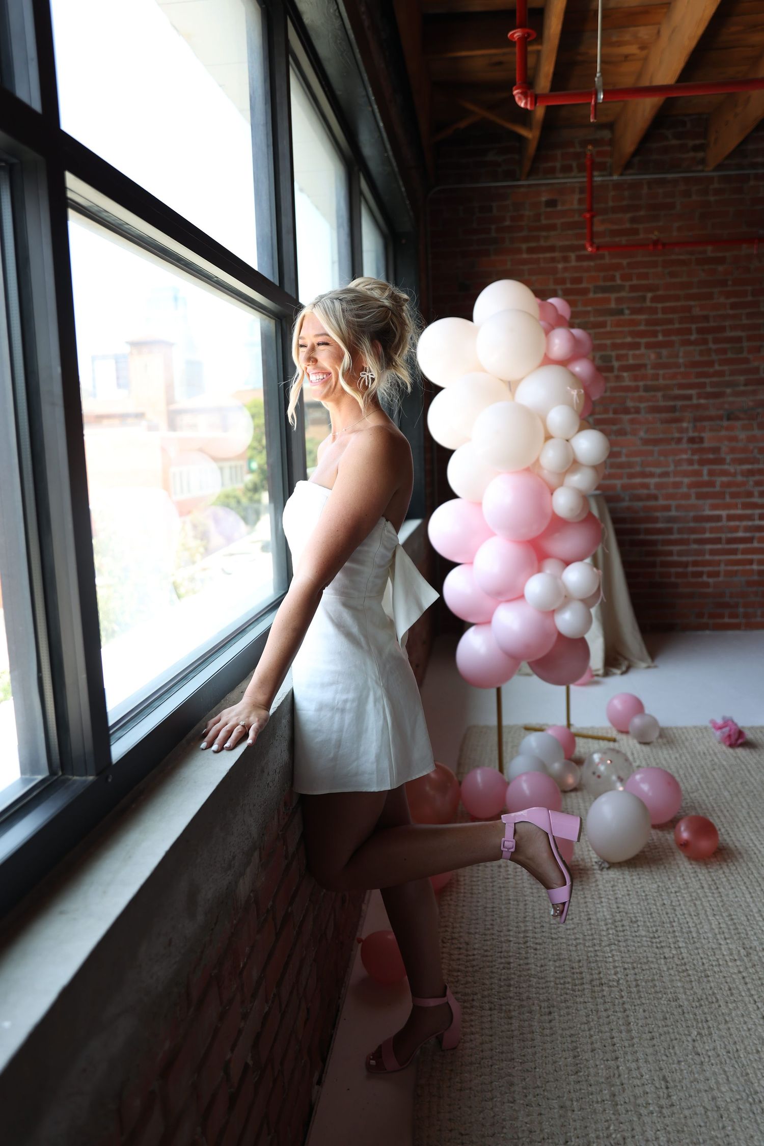 Woman in white dress leans against industrial window with pink balloon decor.