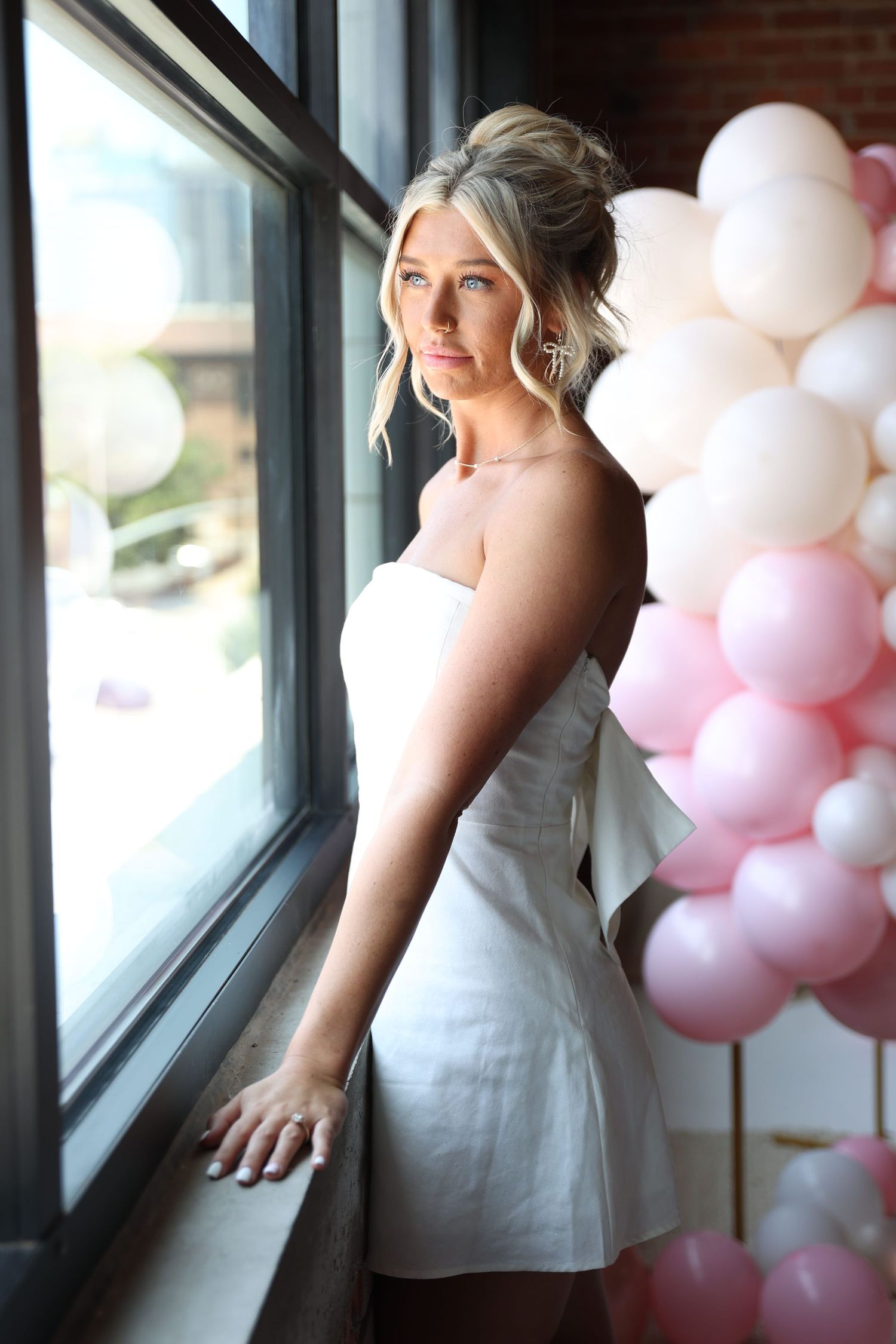 Woman in white dress gazes thoughtfully out window with pink balloon arch in background.