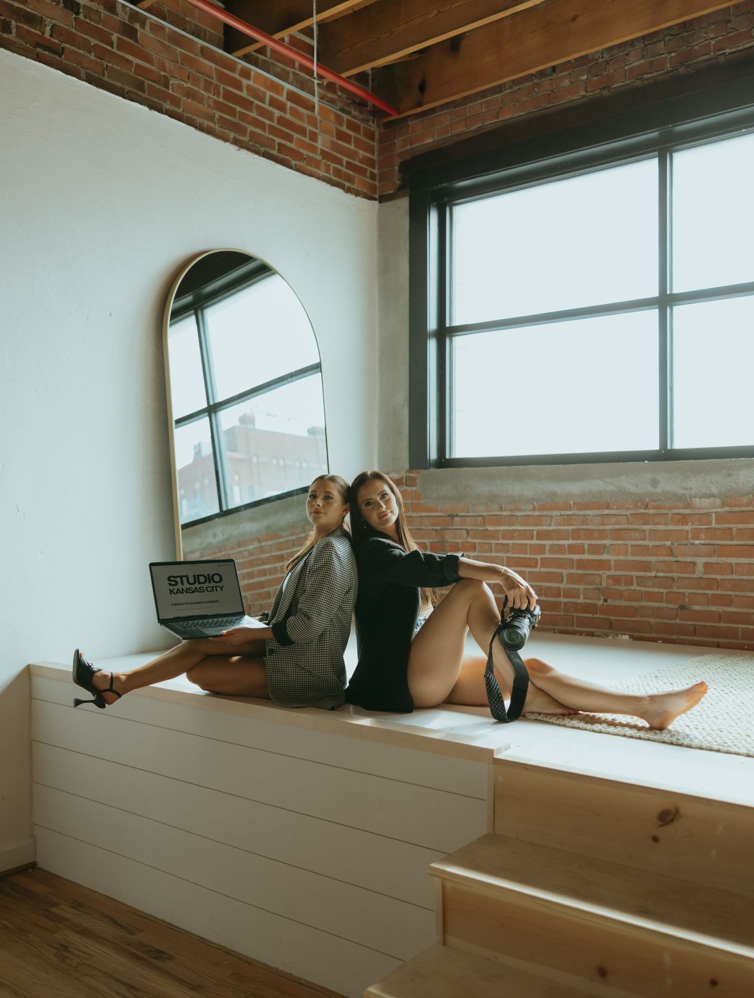 Two people posing back-to-back on a window ledge in an industrial studio setting.