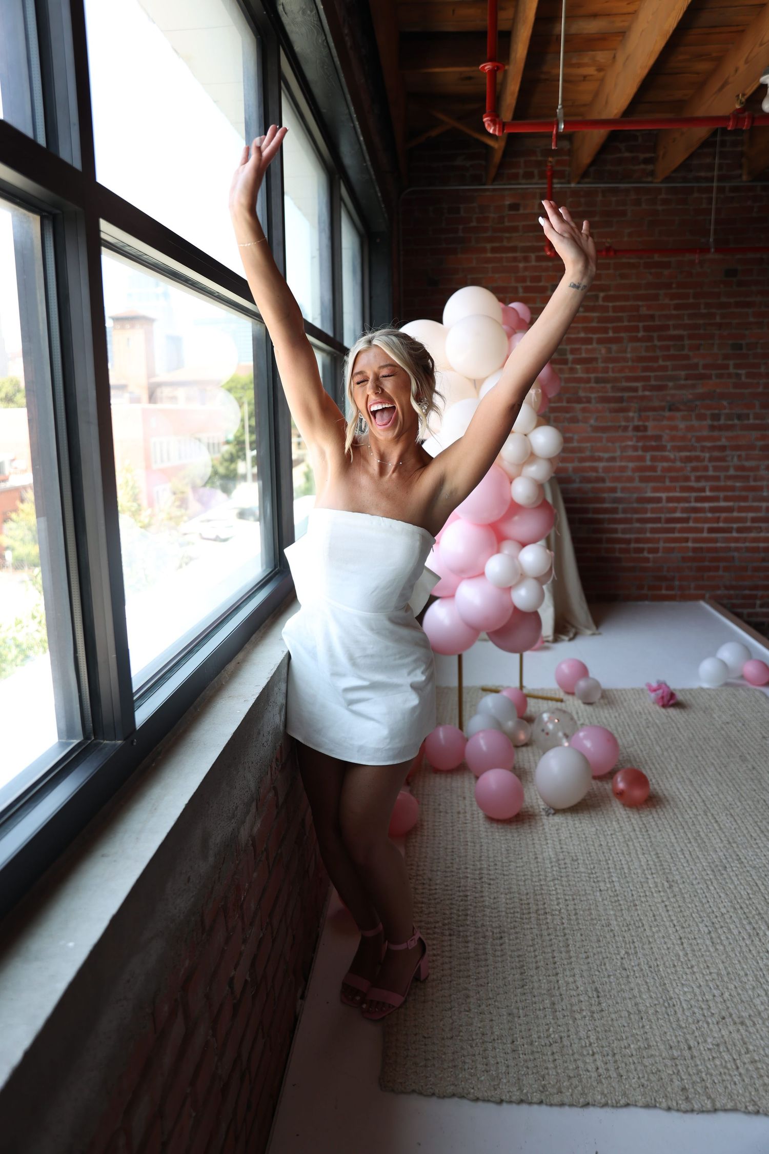 Joyful bride in white dress celebrating with pink and white balloons by industrial window.