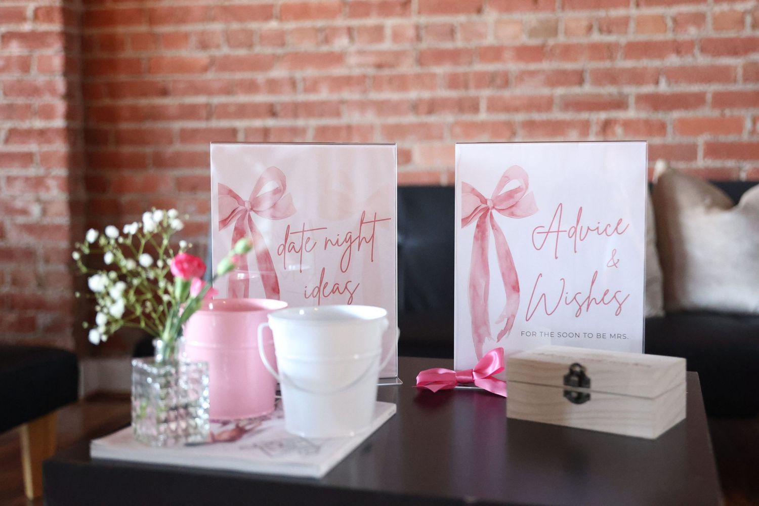 Pink and white party decor setup with signage and floral arrangements against exposed brick.