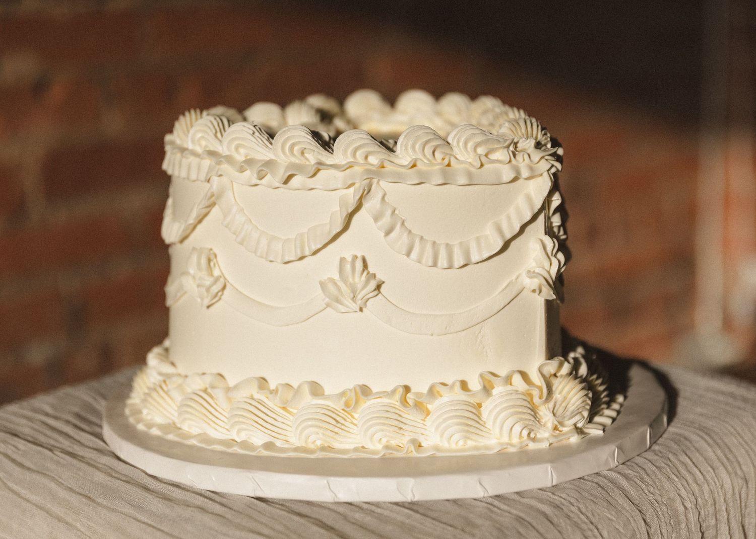 White wedding cake with delicate piped details and scalloped decorative patterns against a brick wall.