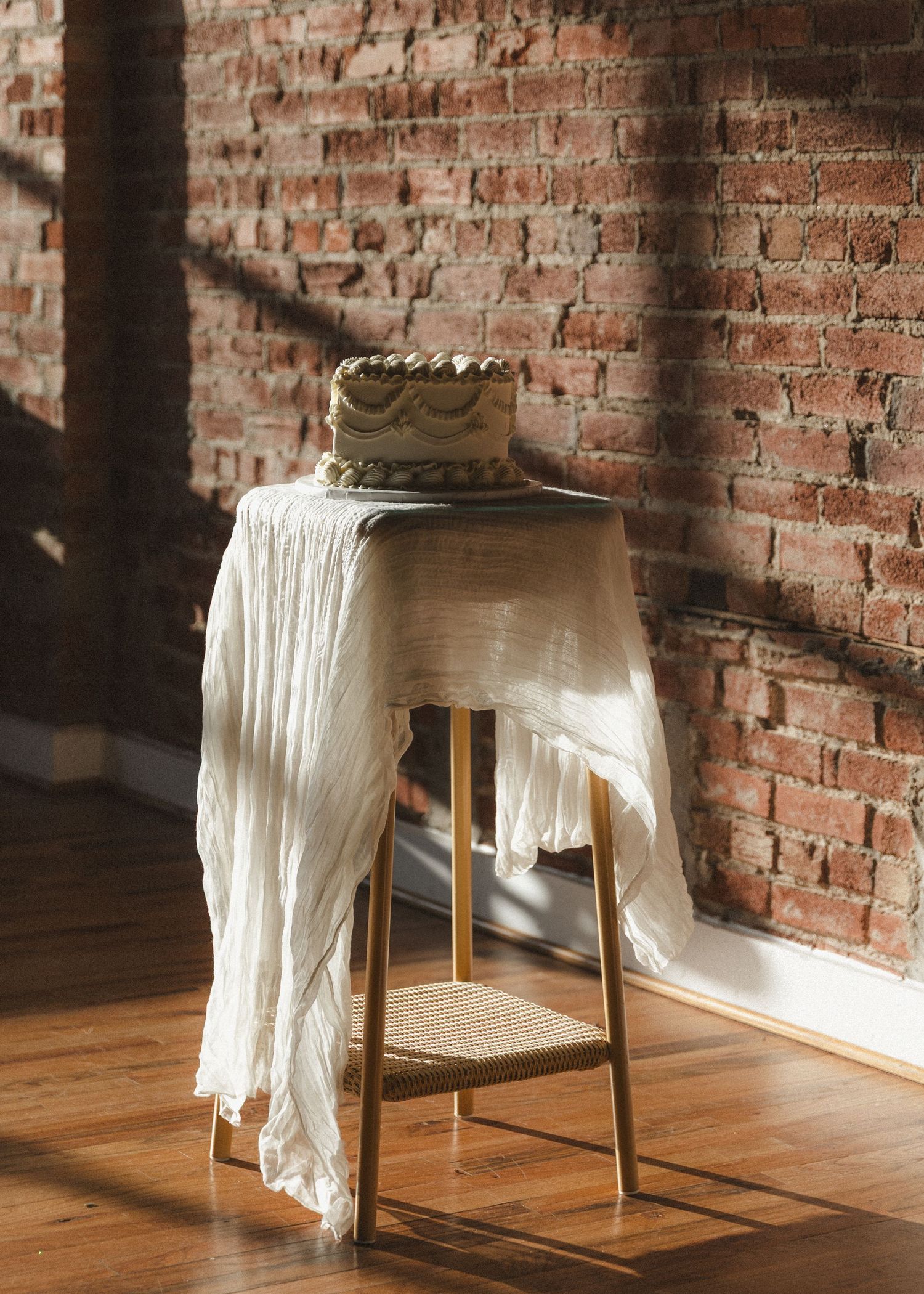 Wedding cake displayed on vintage stool with flowing white fabric against exposed brick wall.
