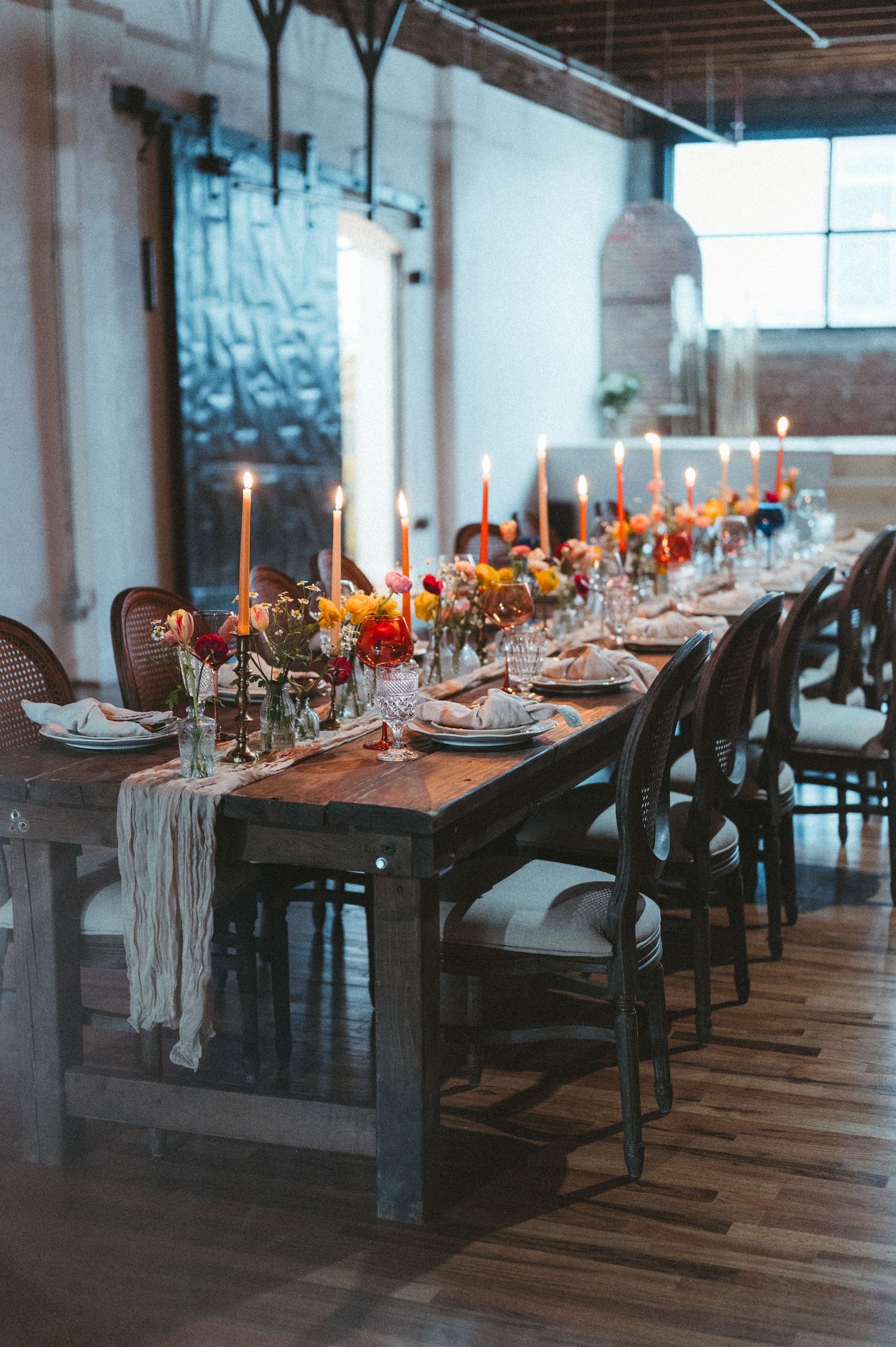 Long wooden dining table decorated with candles and draped fabric in an industrial-style space.
