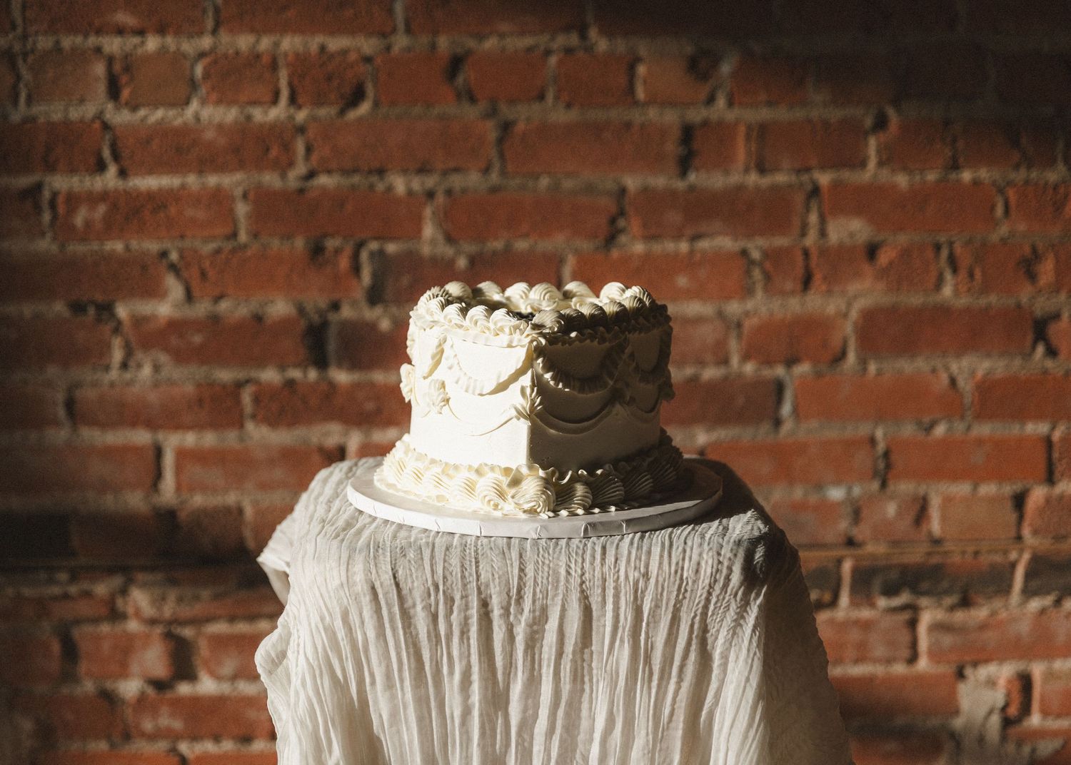 White wedding cake with intricate frosting detail displayed on draped fabric against brick wall.