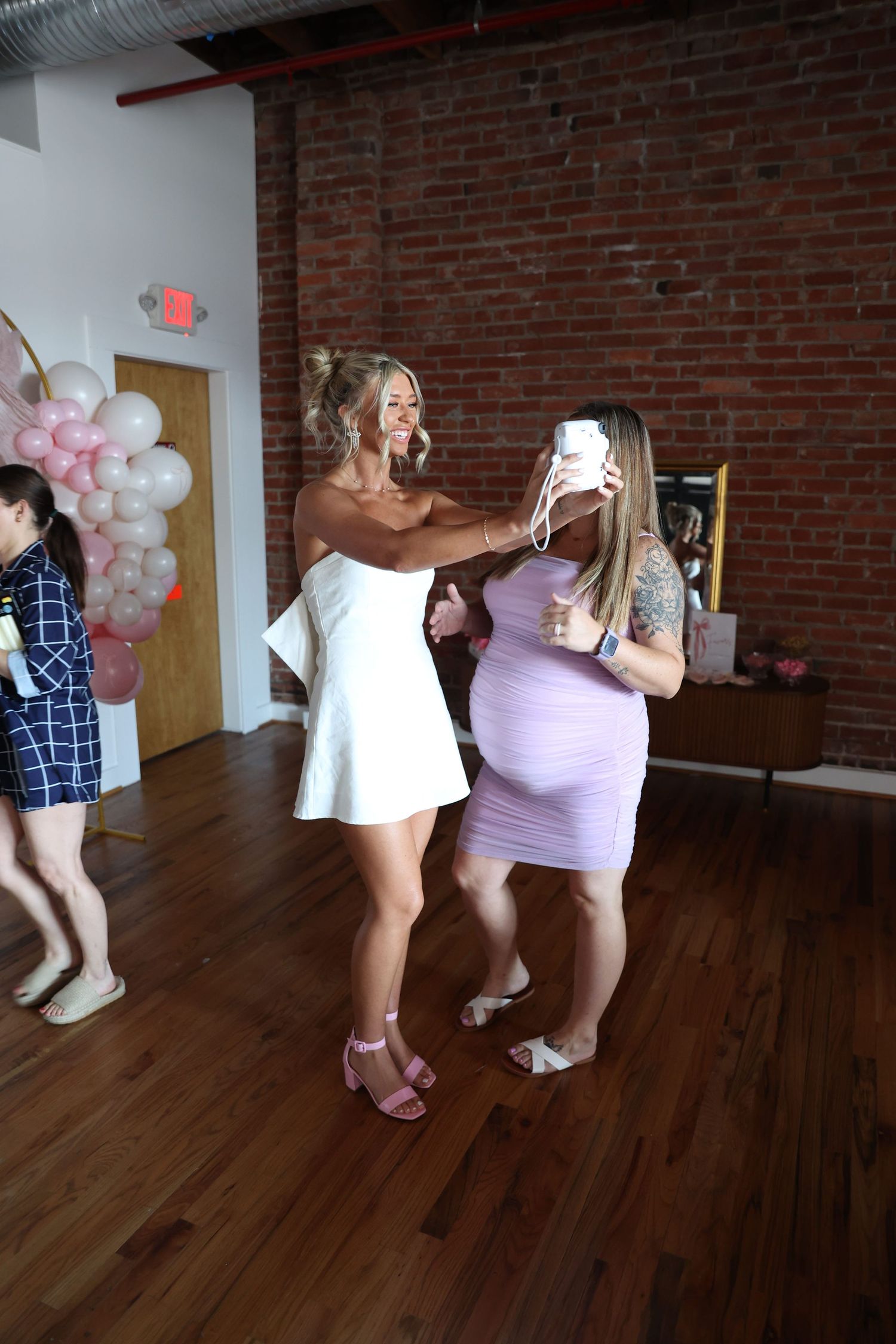 Party guests enjoying a moment at an event with pink balloon decorations and brick wall backdrop.