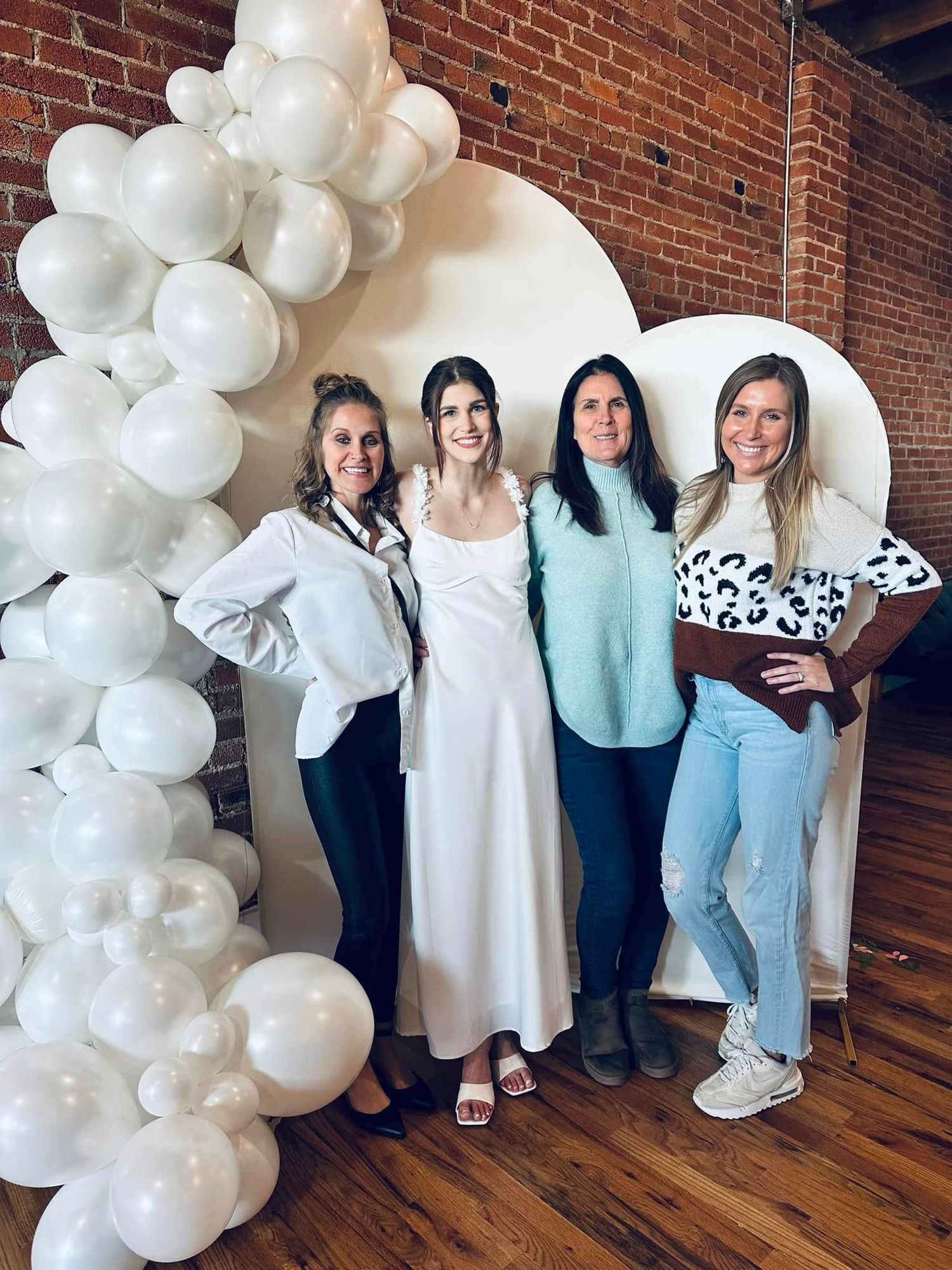 Group photo of four women under white balloon arch in industrial brick venue.