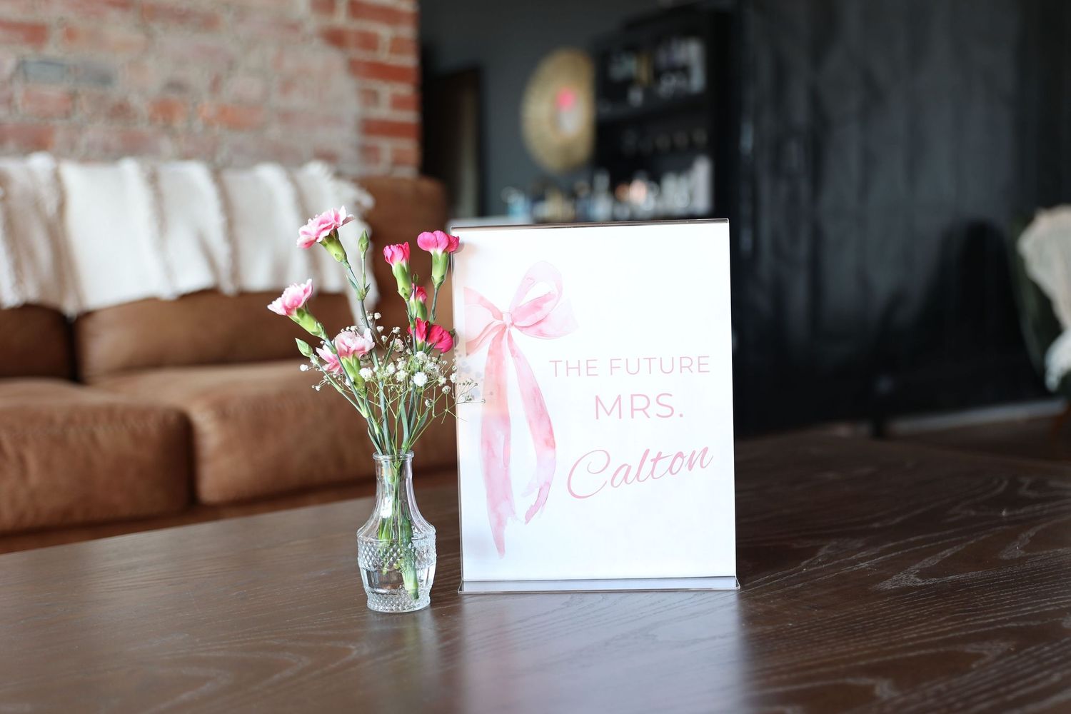 White photo card with pink bow design beside glass vase with pink roses on wooden table.