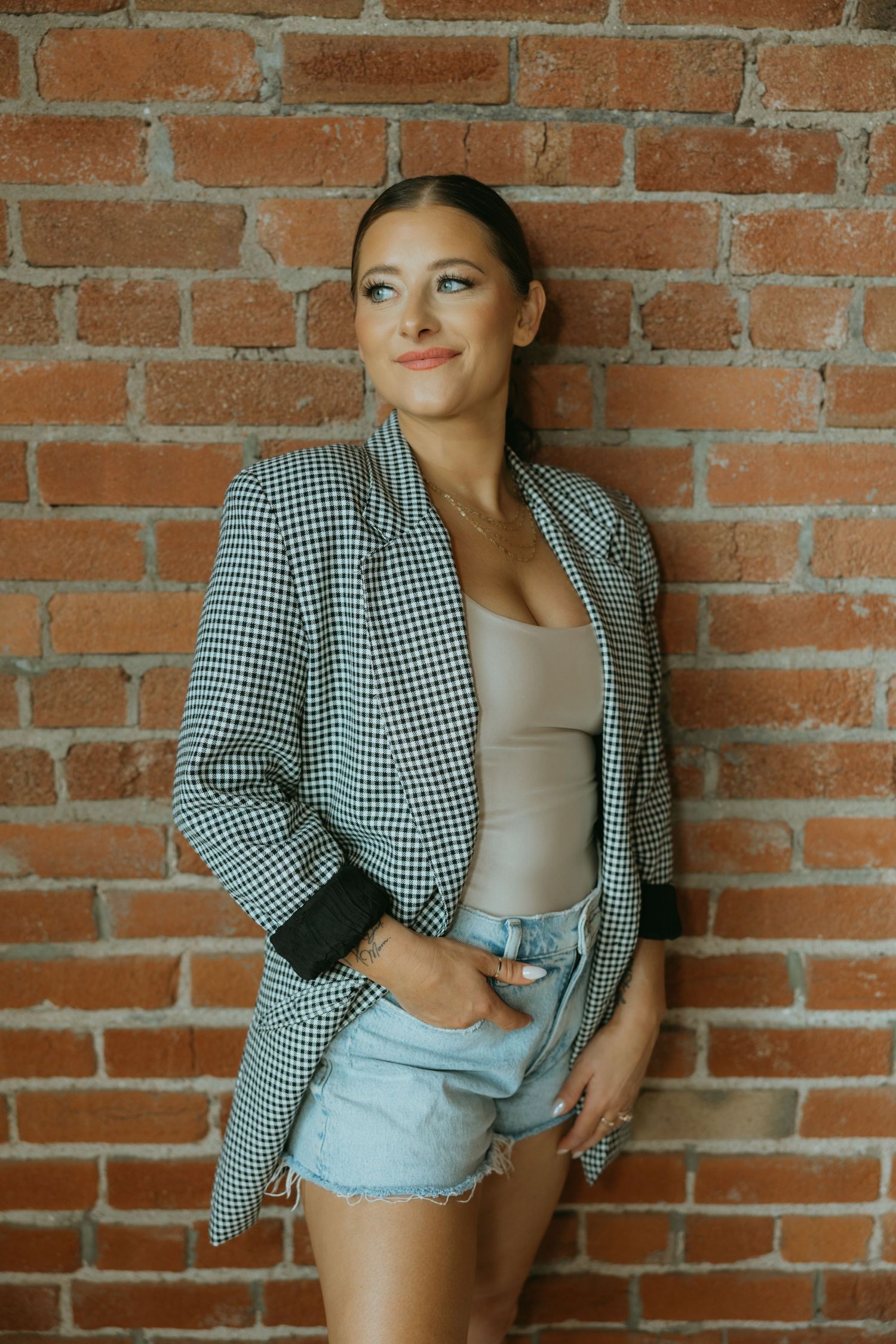 Casual chic look with gingham blazer and denim shorts against brick wall.