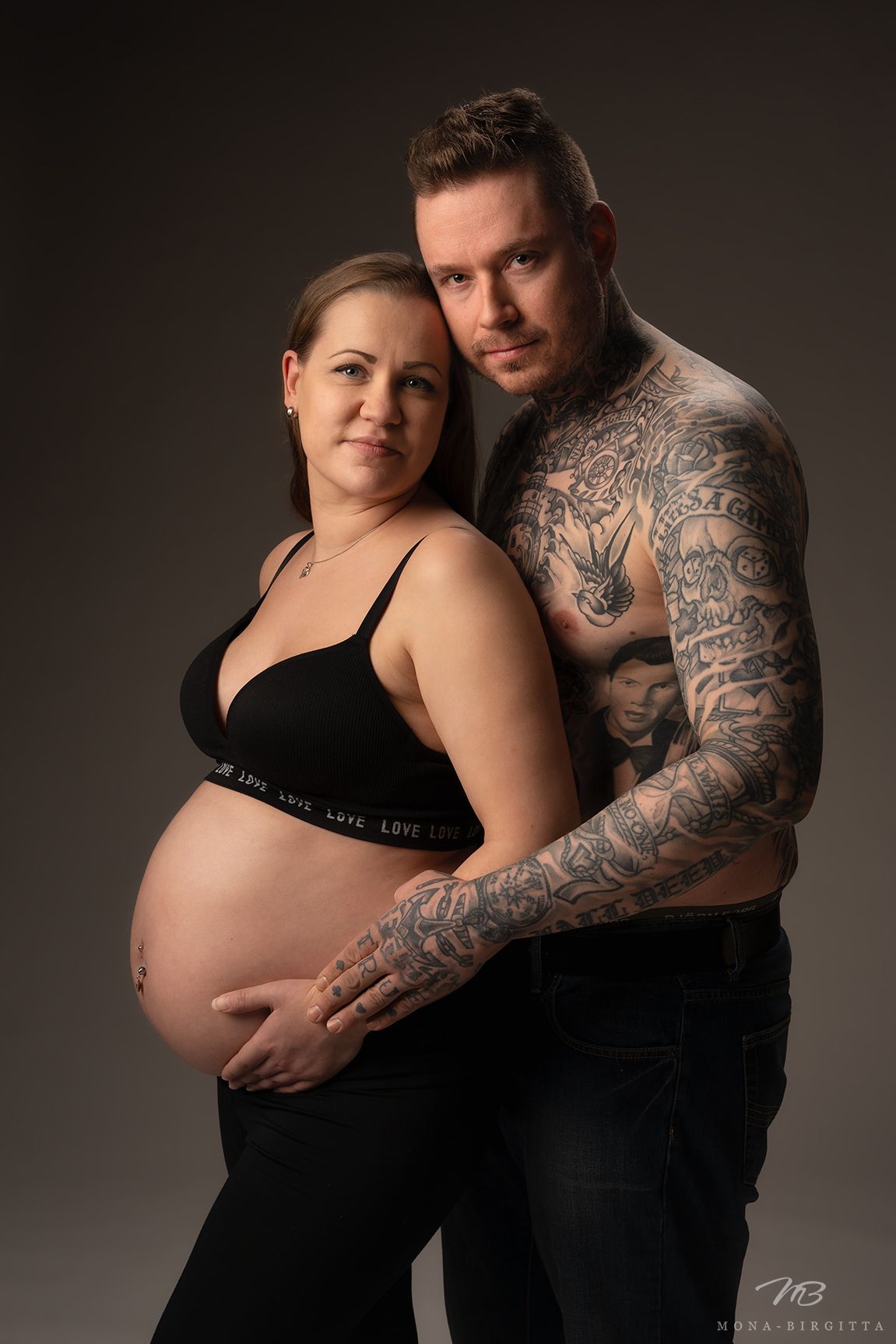 Tatuoitu mies ja raskaana oleva nainen herkässä yhteiskuvassa. Pariskunta pitää molemmat hellästi raskausvatsasta kiinni.