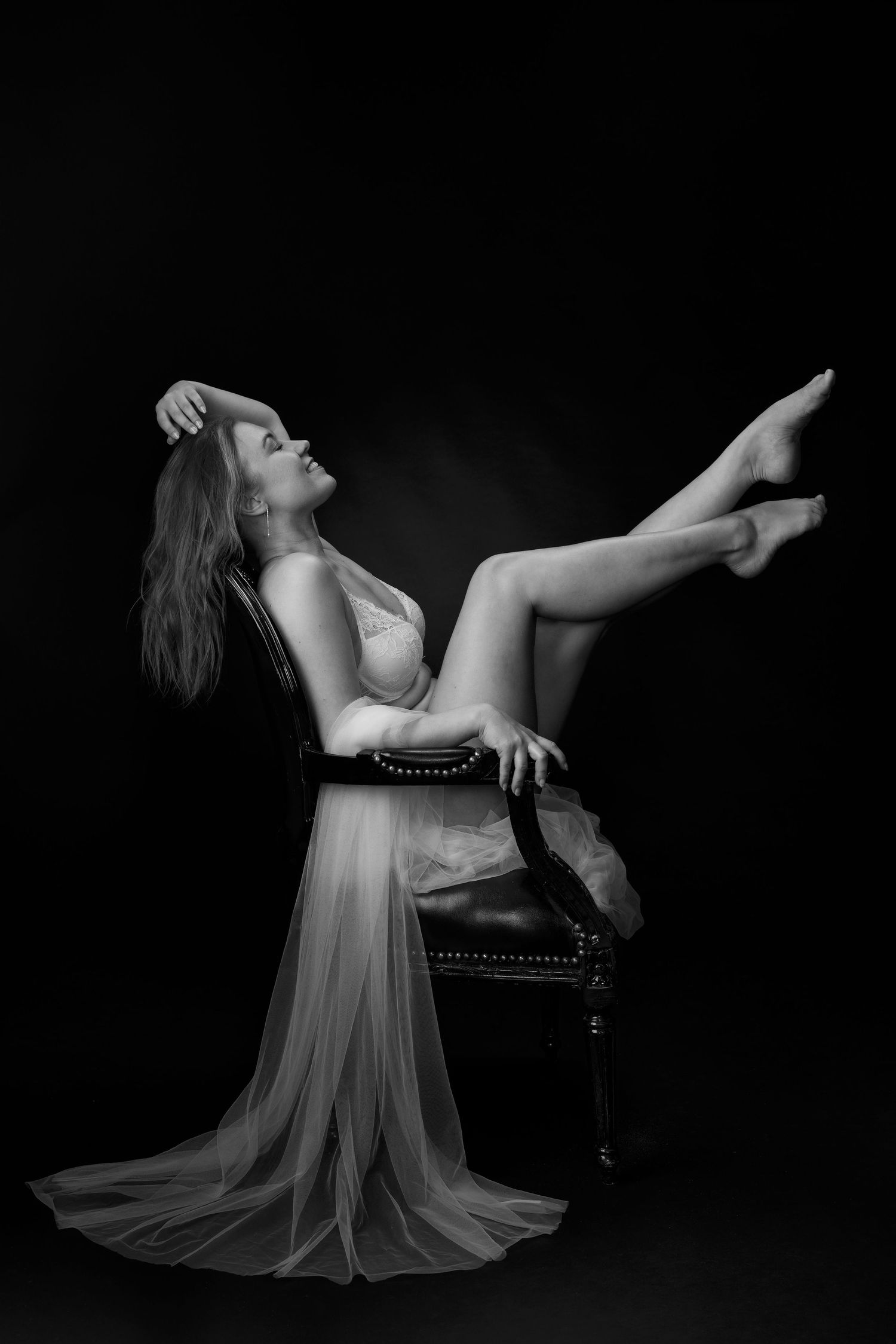 Black and white photo of a woman in a sheer gown posed gracefully on a vintage chair with legs raised against dark background.