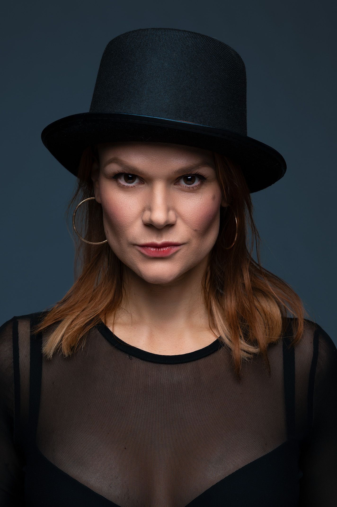 Sinistä taustaa vasten kuvattu muotokuva tummahiuksisesta naisesta jolla on päässään musta silinterihattu ja korvissa kultaiset korvakorut.
